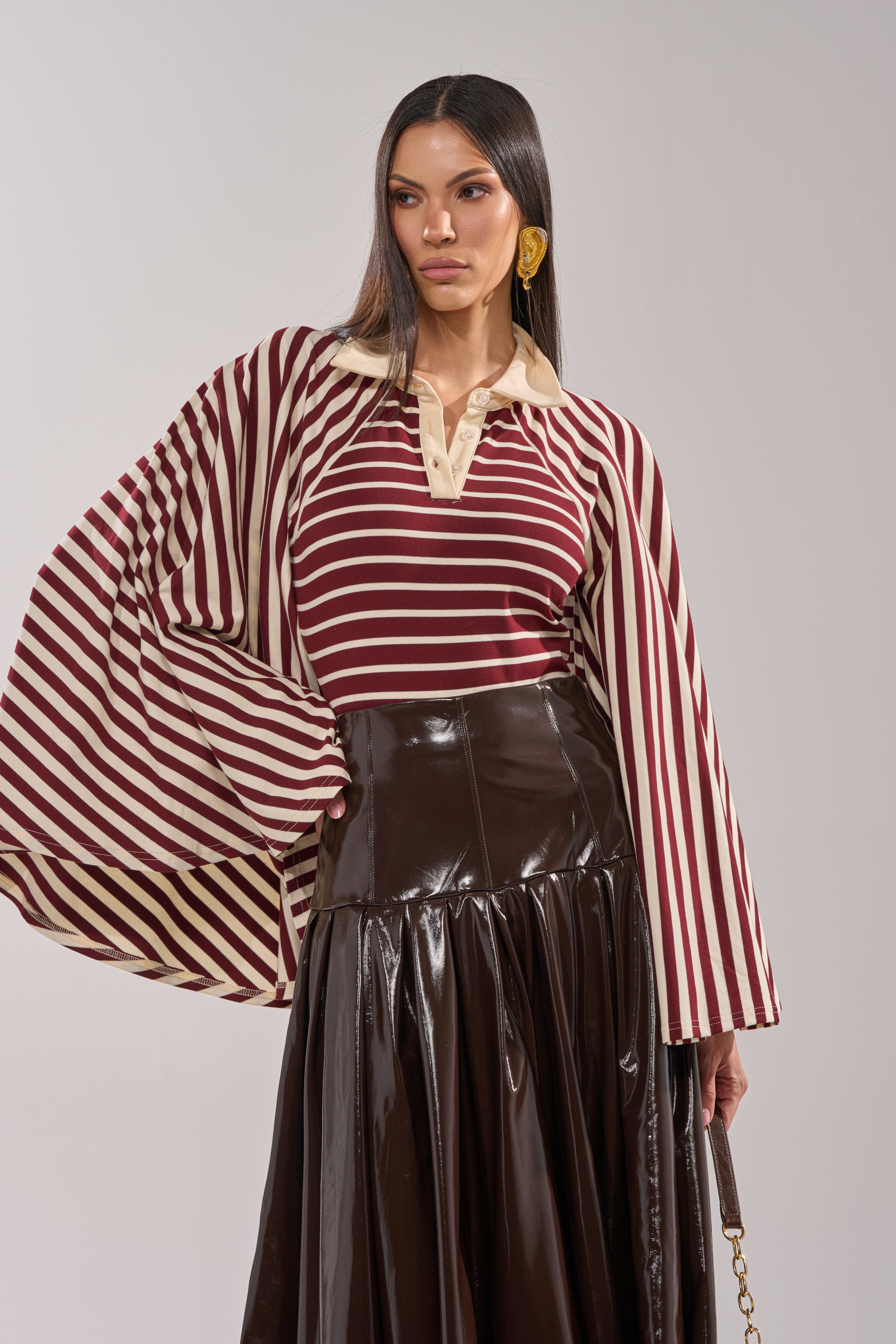 A woman in the ALMA STRIPE COLLARED BODYSUIT with maroon and cream bell sleeves, a dark brown shiny pleated skirt, and large gold earrings stands against a plain light background, gazing to the side with one hand on her hip. 0