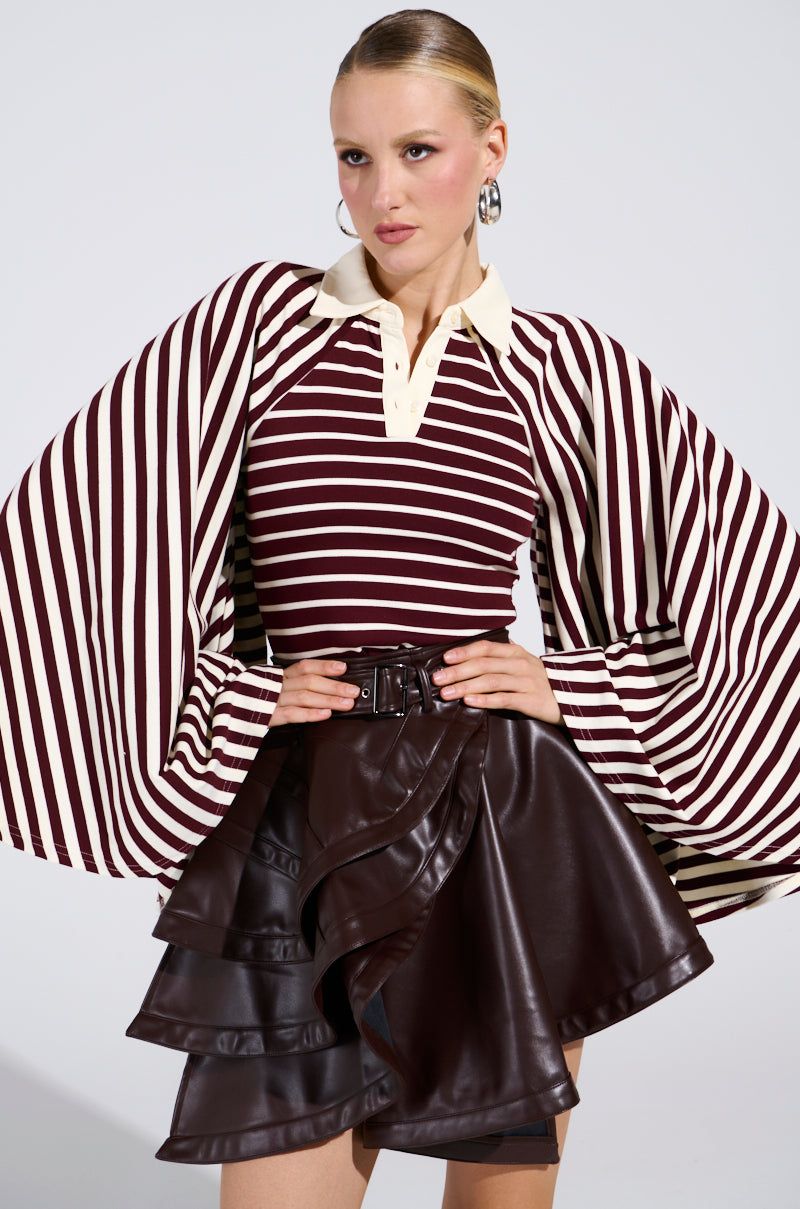 A woman showcases the ALMA STRIPE COLLARED BODYSUIT—maroon and white stripes with a collared neck and dramatic bell sleeves—paired with a dark brown ruffled leather mini skirt and hoop earrings, posed against a plain light background.