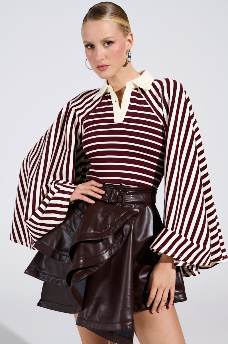 A person models the ALMA STRIPE COLLARED BODYSUIT, a vintage-inspired maroon and white striped piece with a collar and bell sleeves, styled with a high-waisted dark brown faux leather ruffled skirt and hoop earrings.