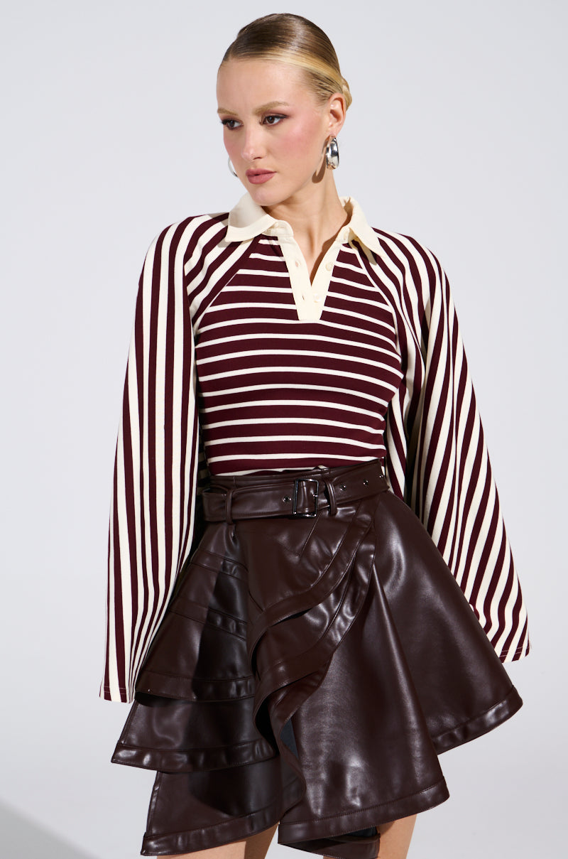 A woman wears the ALMA STRIPE COLLARED BODYSUIT in maroon and cream stripes, paired with a high-waisted dark brown ruffled leather skirt. Slicked-back hair and bold earrings complete her modern yet timeless style.