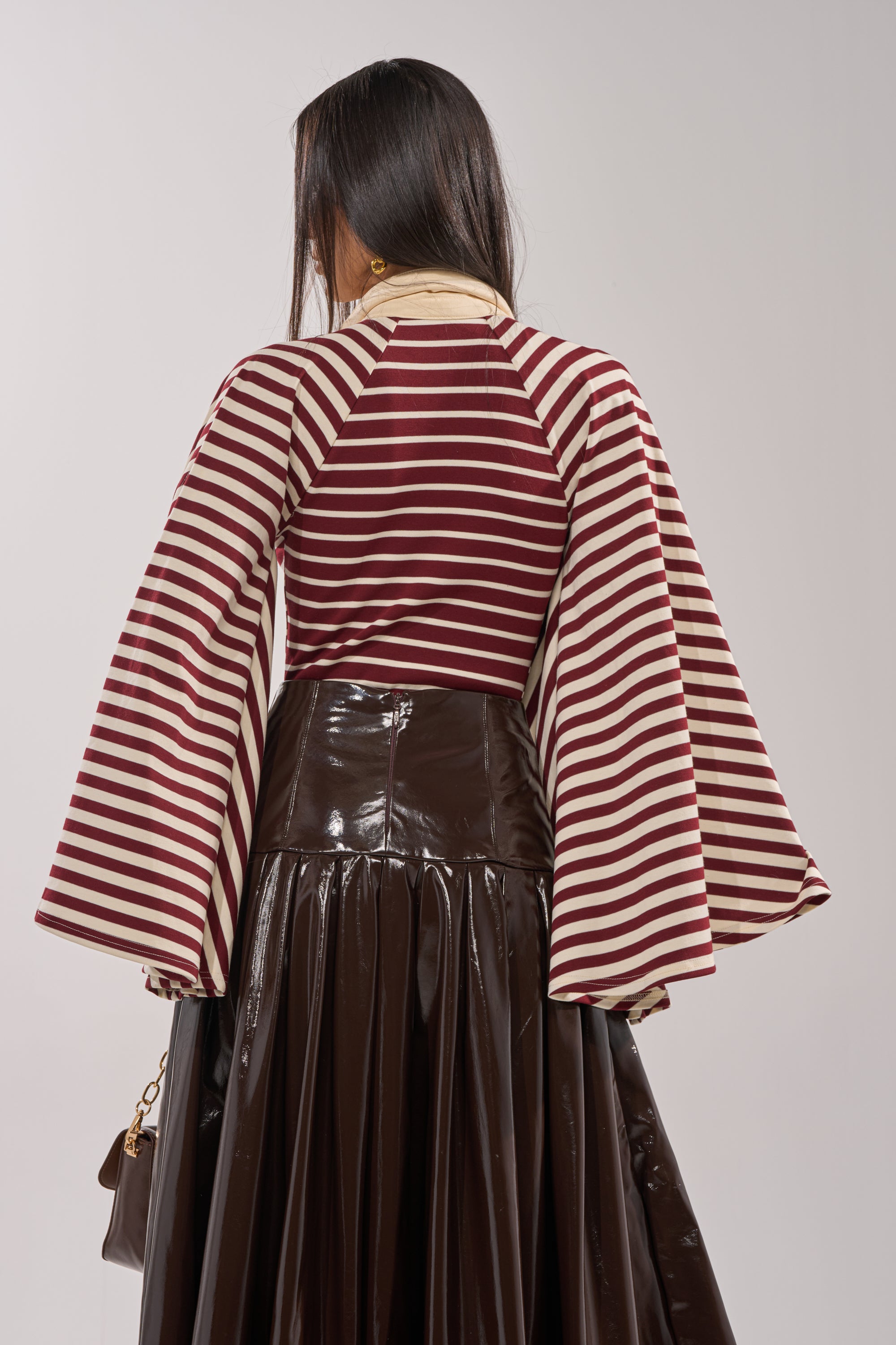 A person with long dark hair, seen from behind, wears the ALMA STRIPE COLLARED BODYSUIT in maroon and white stripes with wide sleeves, paired with a shiny dark brown pleated skirt and holding a small brown handbag. 4