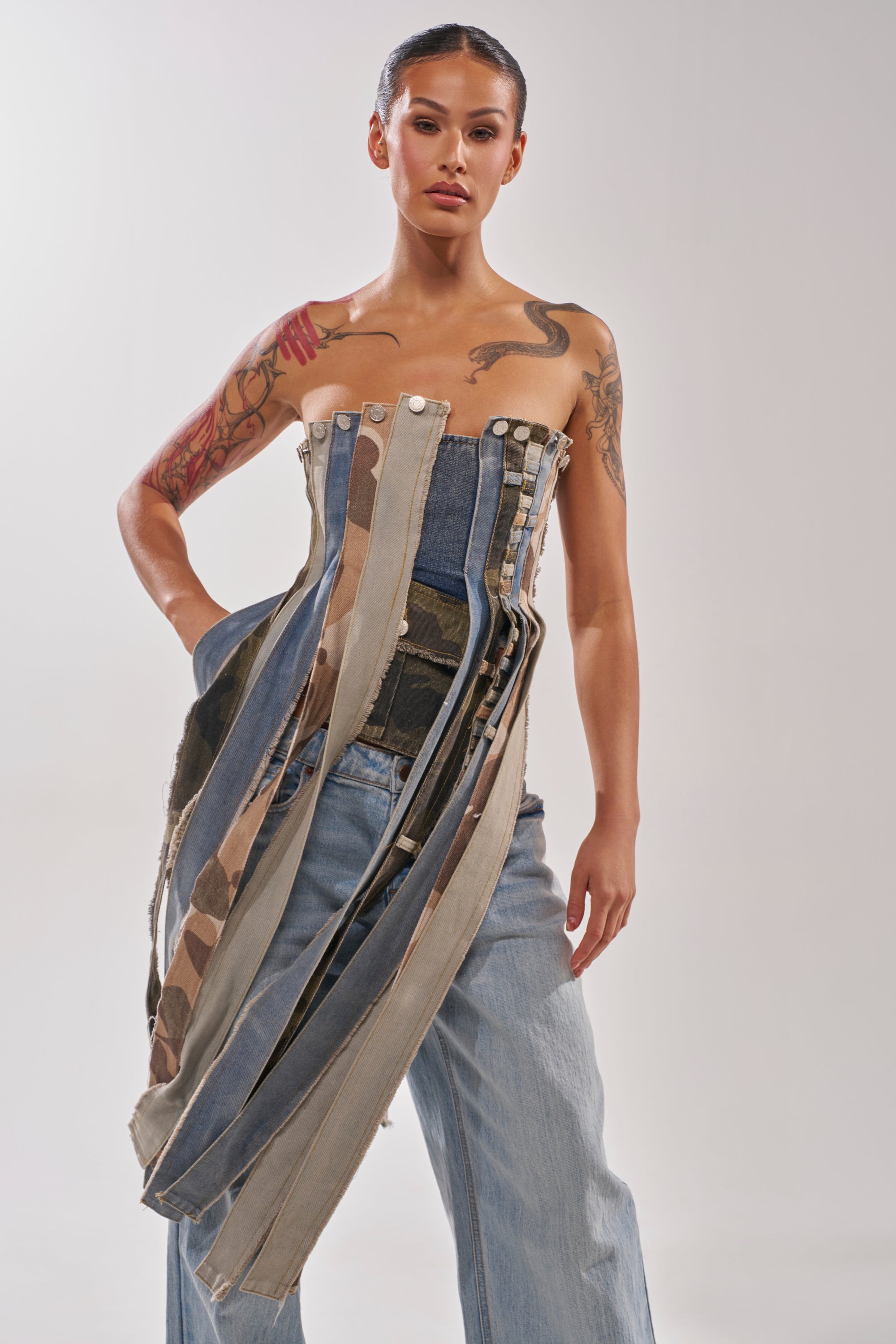 A tattooed woman stands confidently against a plain background wearing the MIXED EMOTION CORSET—a mixed-denim, vertically striped corset top—paired with light blue jeans. 0