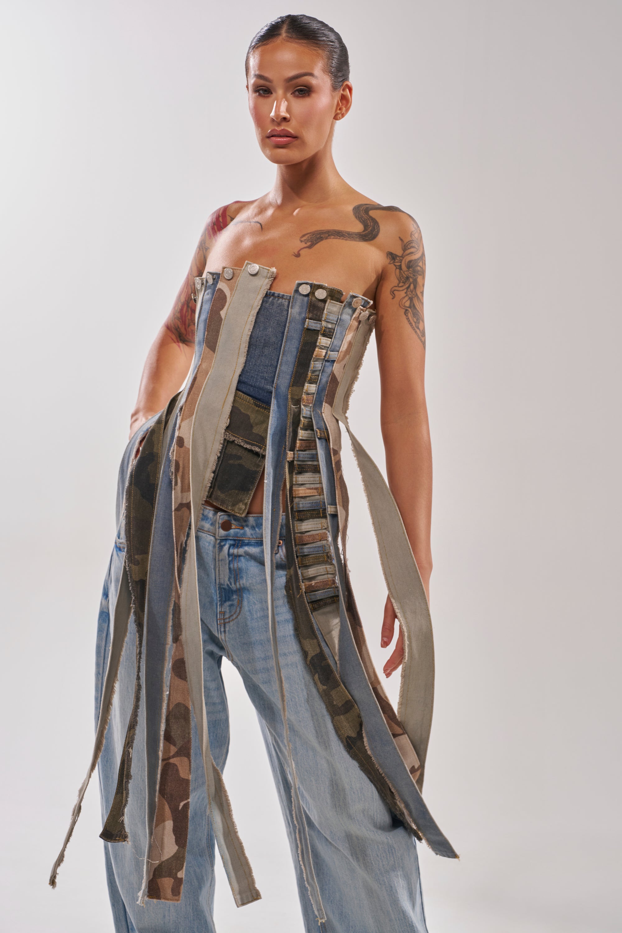 A tattooed woman stands confidently in the MIXED EMOTION CORSET, a unique denim top with fringe and camouflage details, paired with loose light blue jeans against a plain background. 1