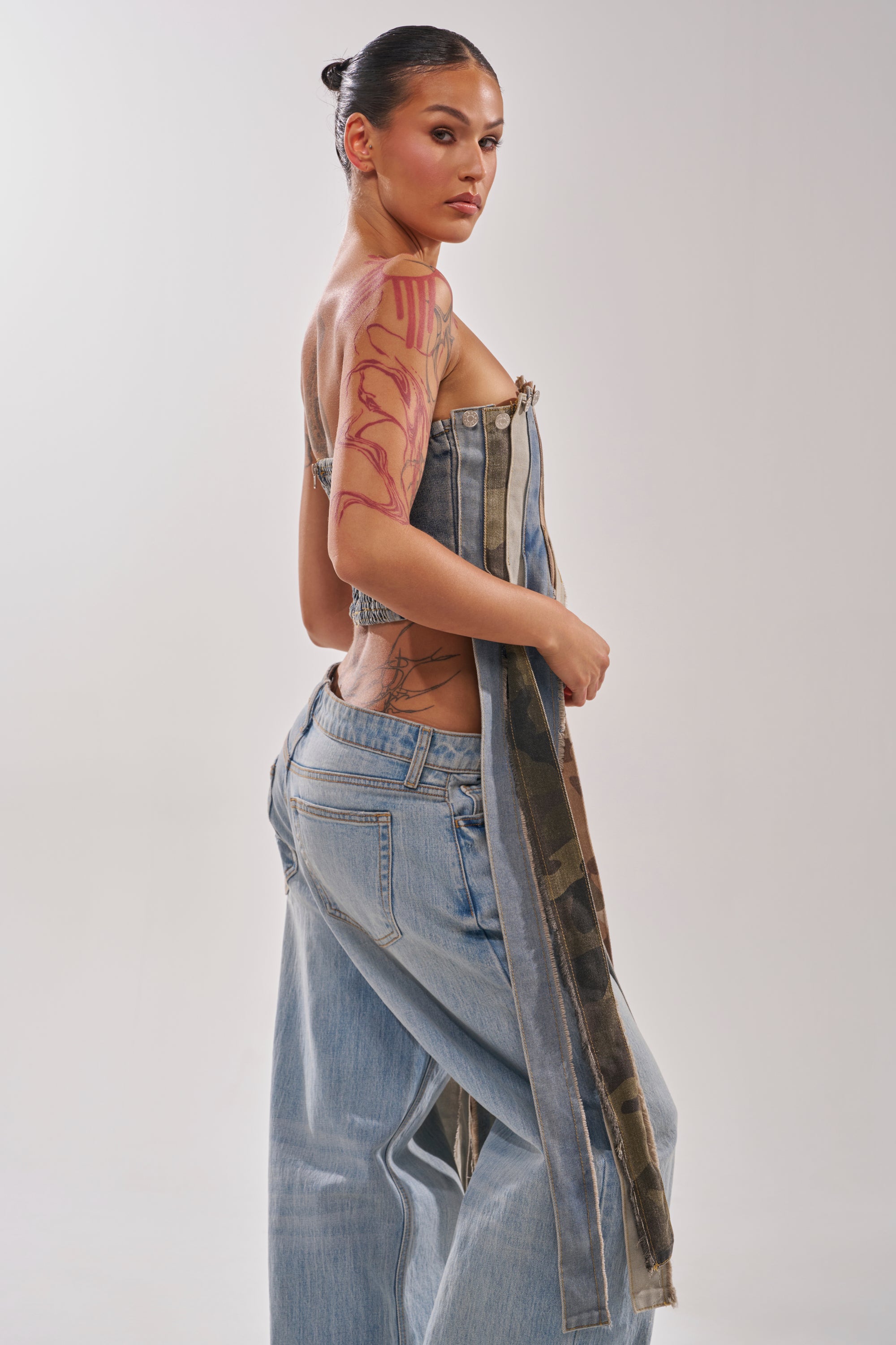 A woman with slicked-back hair and visible tattoos models the MIXED EMOTION CORSET, a unique deconstructed denim top, paired with loose light blue jeans. She looks over her shoulder against a plain background. 2