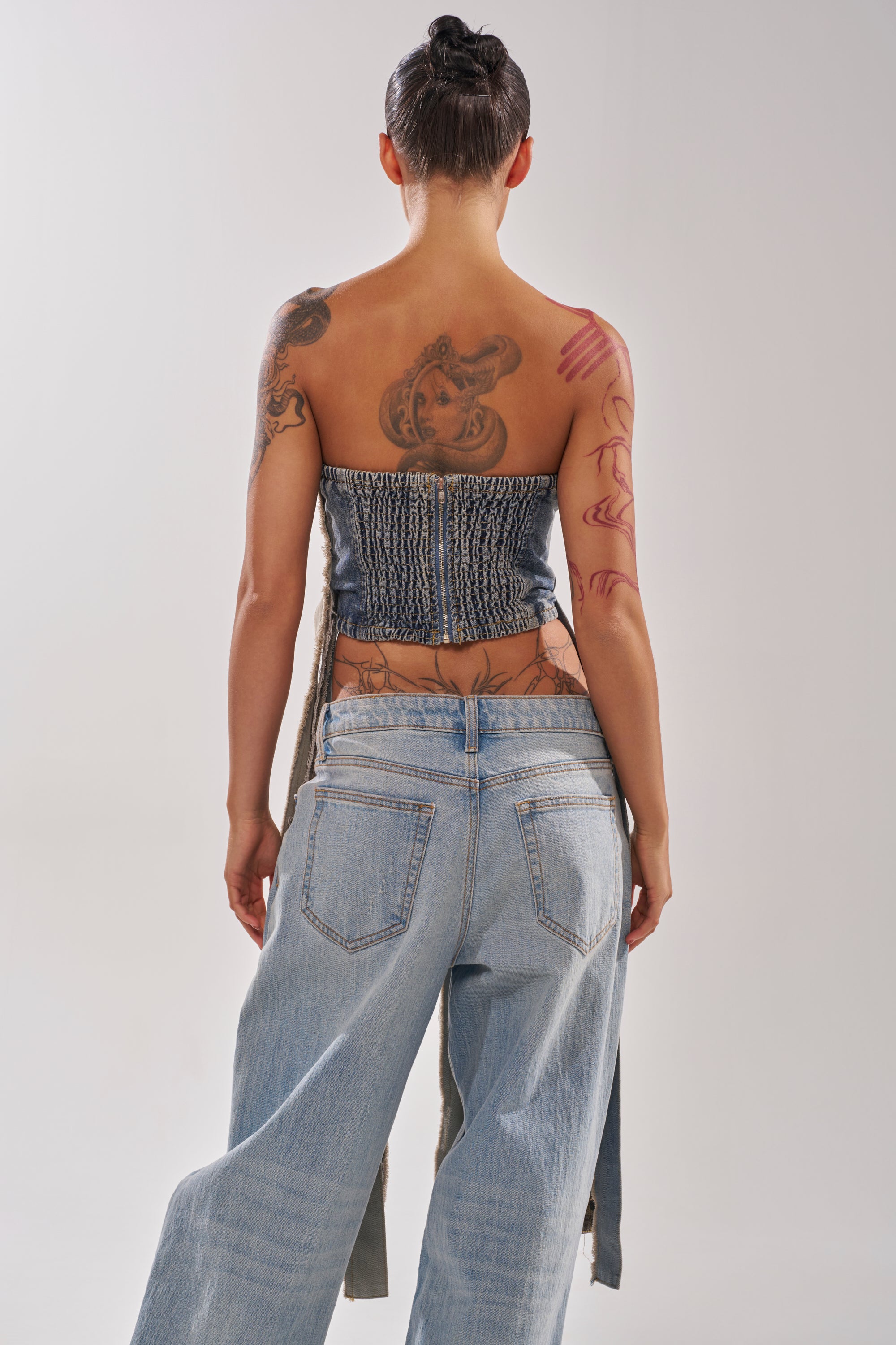 A person with tattoos on their arms and back stands facing away, wearing the strapless MIXED EMOTION CORSET and loose-fitting light blue jeans against a plain, light background. 3