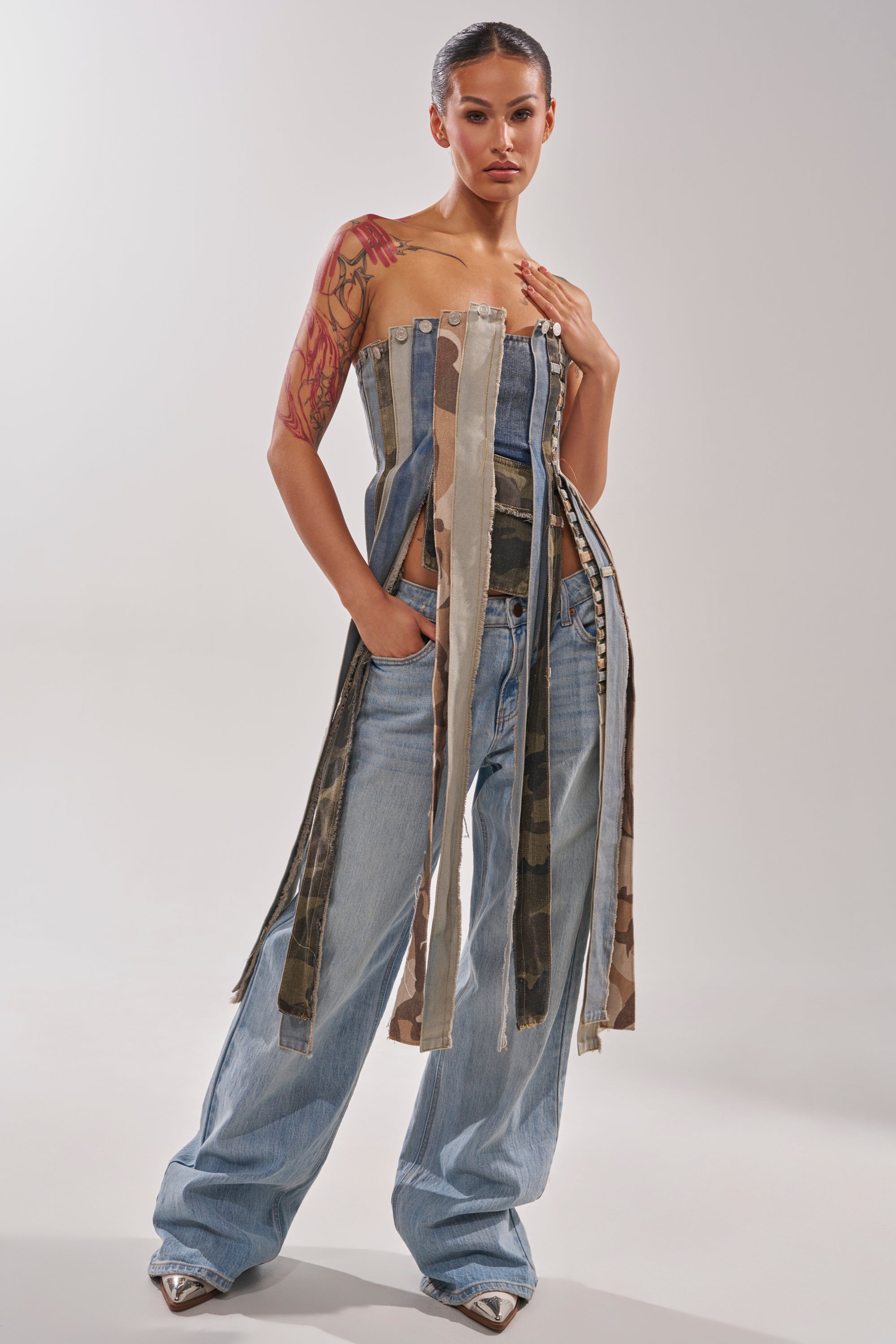A woman poses confidently in light blue wide-leg jeans and the MIXED EMOTION CORSET—a strapless denim piece with long, multicolored fabric strips. She has slicked-back hair, visible tattoos, and silver pointed-toe shoes. 4