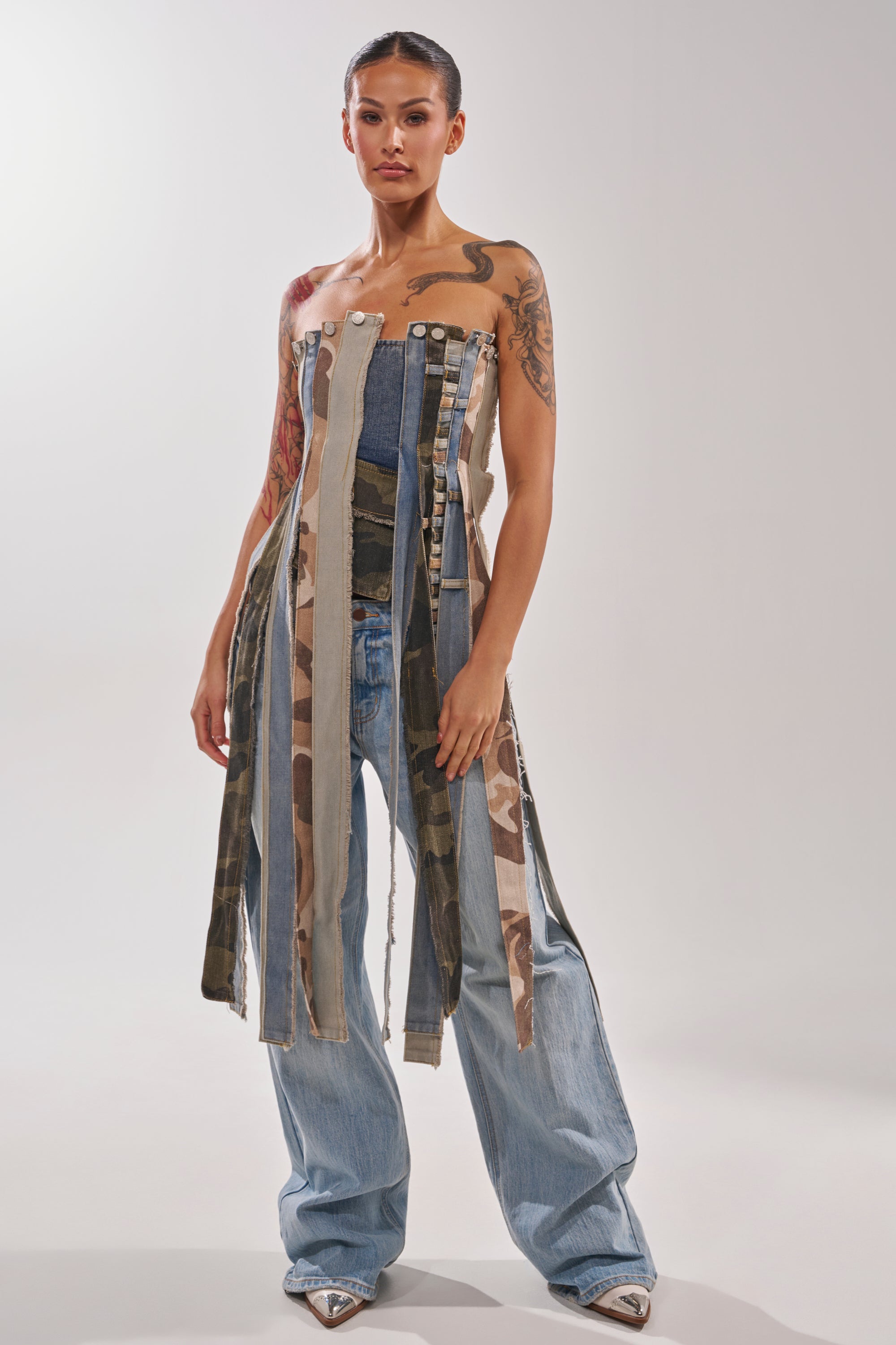 A woman with slicked-back hair and tattoos faces forward, wearing wide-leg jeans and the MIXED EMOTION CORSET—a bold deconstructed denim and camouflage top with long strips hanging to her knees. 5