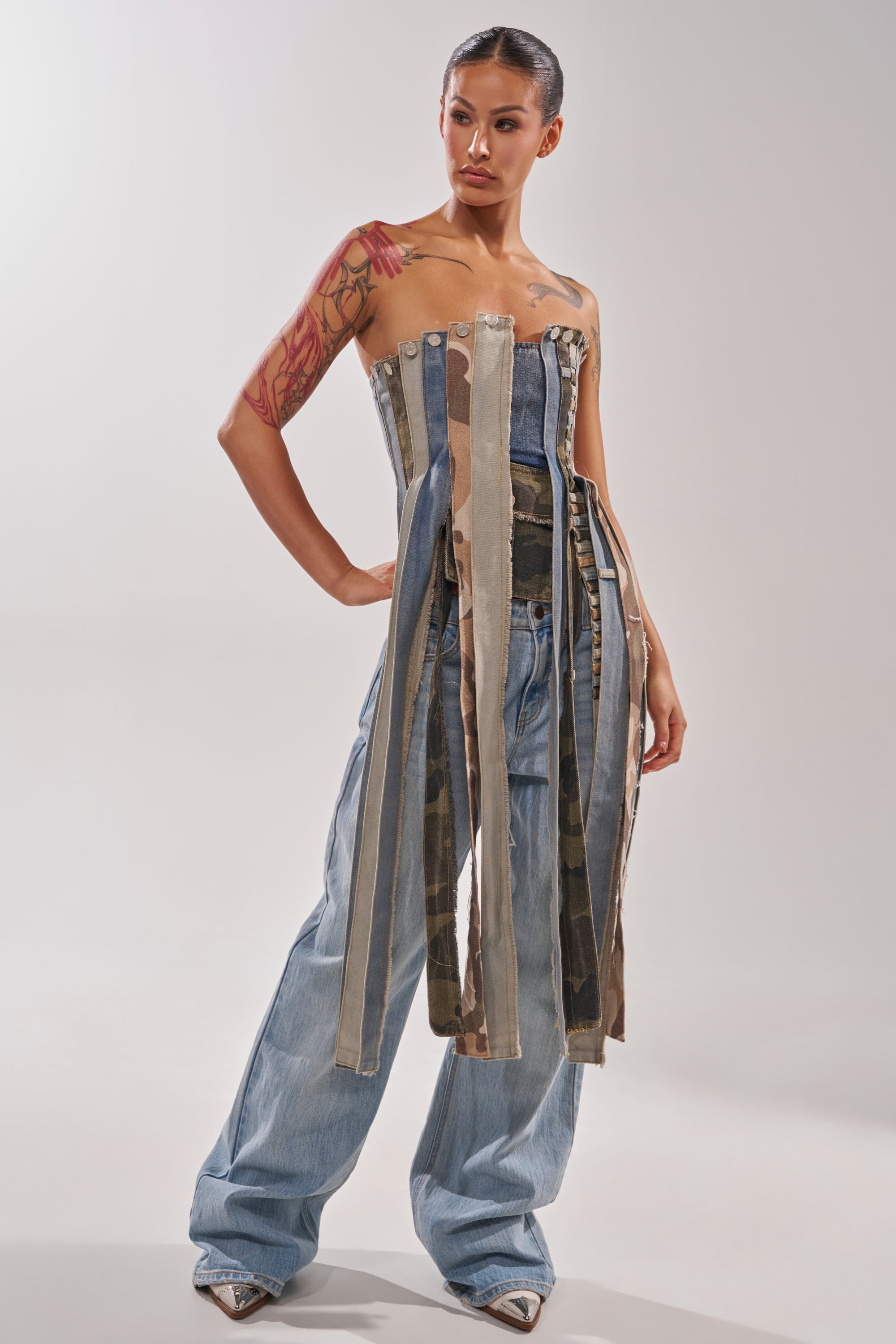 A woman with a tattooed arm poses in the MIXED EMOTION CORSET, a strapless fringe denim top with long hanging strips, styled over loose blue jeans and pointed shoes, against a plain light background. 6