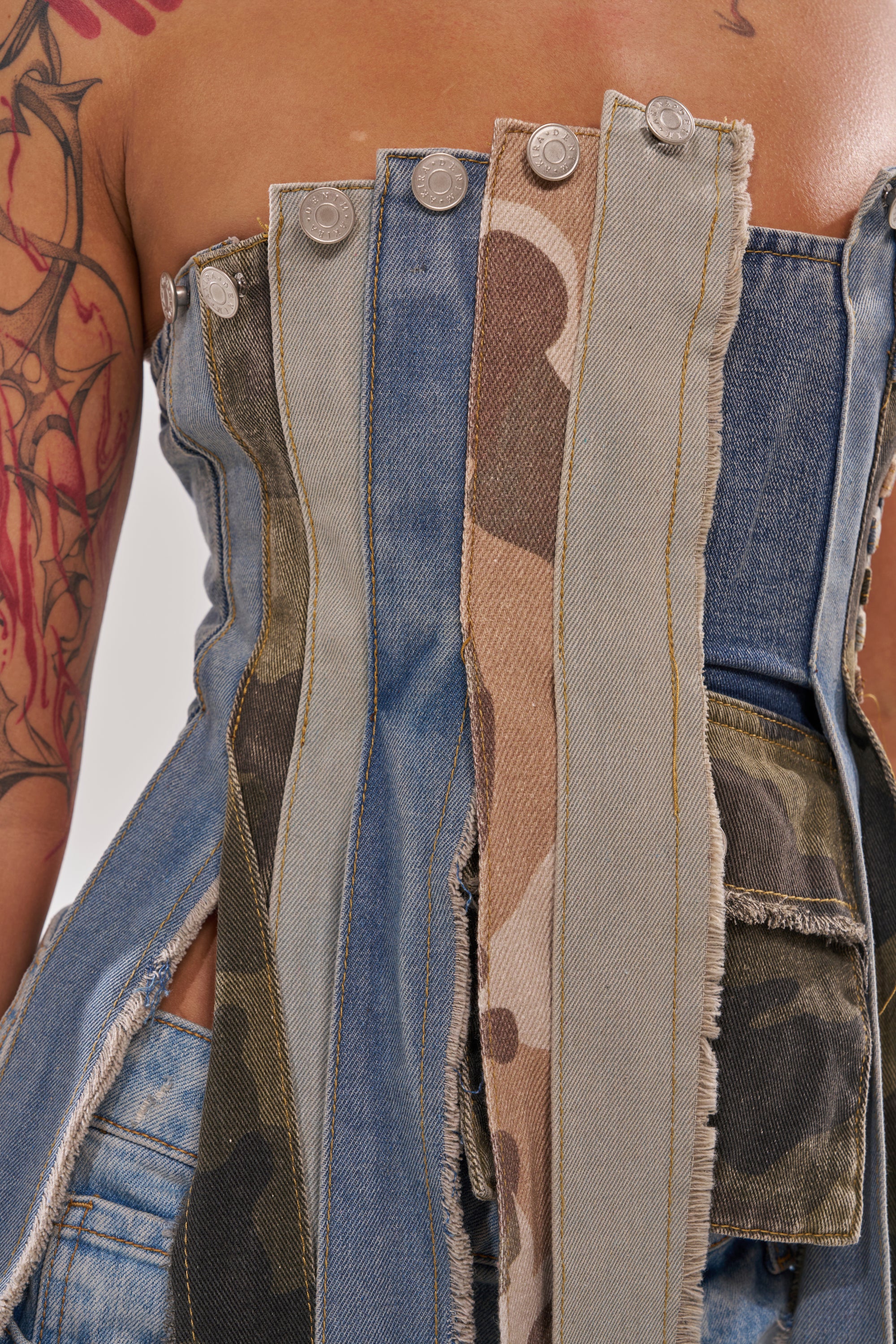 A close-up of the MIXED EMOTION CORSET features alternating denim and camouflage strips with metal buttons; part of a tattooed arm appears on the left. 7