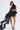 A woman poses against a white background wearing the ROSE PETAL LOVER BLOUSE, a dramatic black sheer tulle top with an oversized rose appliqué on one shoulder, paired with black shorts and strappy black heels.