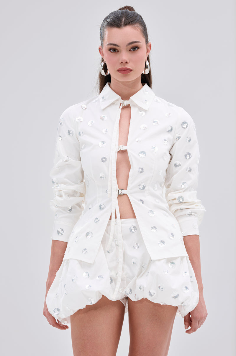 A woman models the LUX FASHION BLOUSE, a white cotton-blend shirt with metallic closures, paired with a floral sequin bubble skirt and hoop earrings, set against a light gray background.