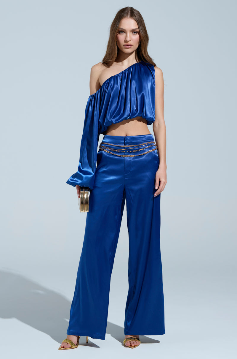 A woman is posing in a shiny blue blouse and silky satin ROYAL FOR LIFE PALAZZO PANTs. She holds a small gold clutch and wears gold heels against a plain, light-colored background.