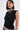 A woman with a bob haircut wears the FOREVER AND EVER SLINKY TOP WITH LACE UP BACK, styled with large hoop earrings and a cross-pendant statement necklace. She has bold eye makeup and stands against a plain light background.