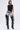 A woman faces forward wearing the FOREVER AND EVER SLINKY TOP WITH LACE UP BACK, distressed denim shorts, and black thigh-high fringe boots. She accessorizes with a bold necklace and hoop earrings.
