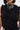 A woman wears the FOREVER AND EVER Slinky Top with lace-up back, featuring ruched ruffle detailing. She accessorizes with a chunky chain necklace adorned with cross pendants, large hoop earrings, and has short, straight hair.