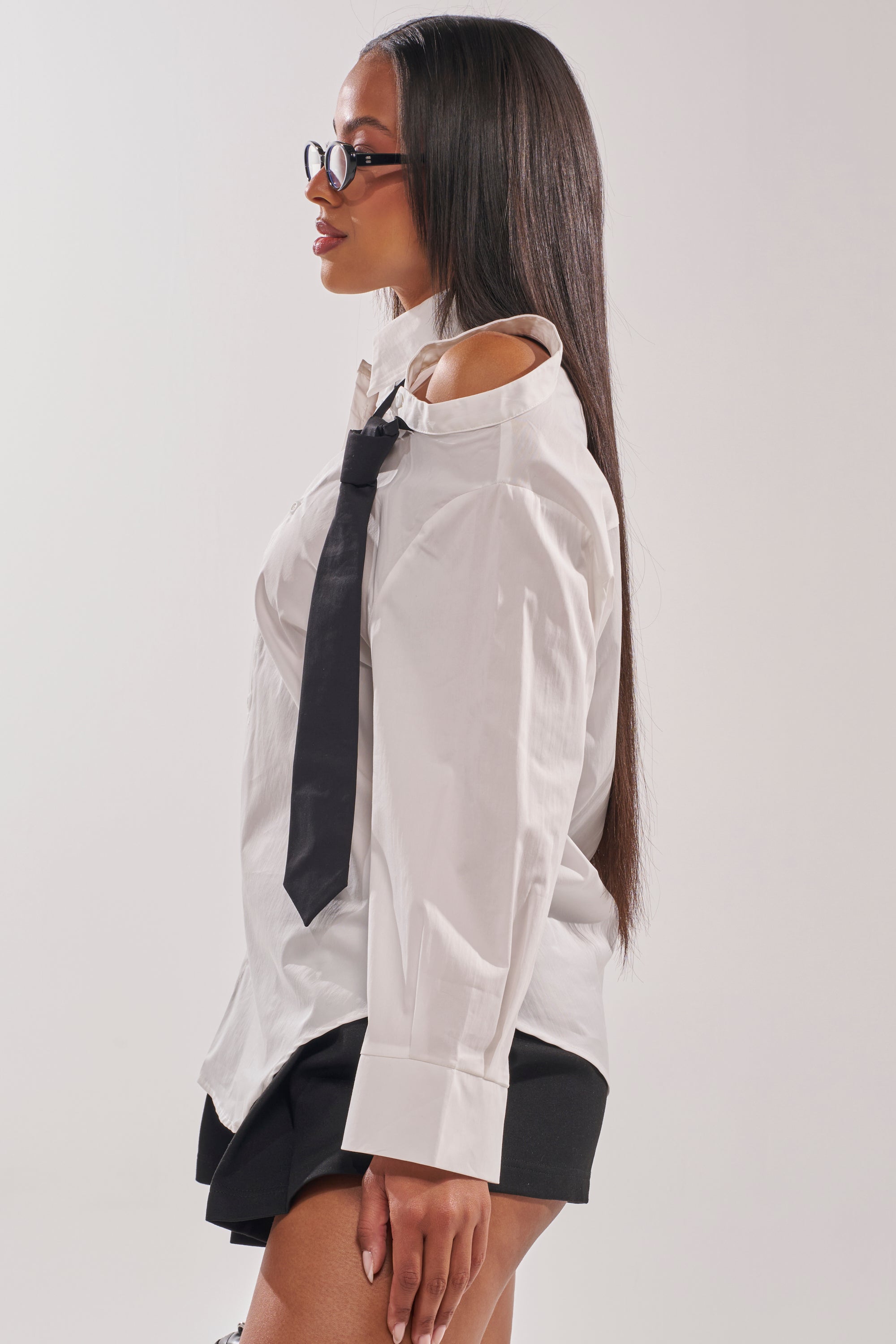 A woman with long straight hair and glasses wears the MIXED UP STRIPED POLO BLOUSE, featuring an asymmetrical white design with open shoulders, paired with a black tie and shorts. She stands in profile against a plain light background. 2