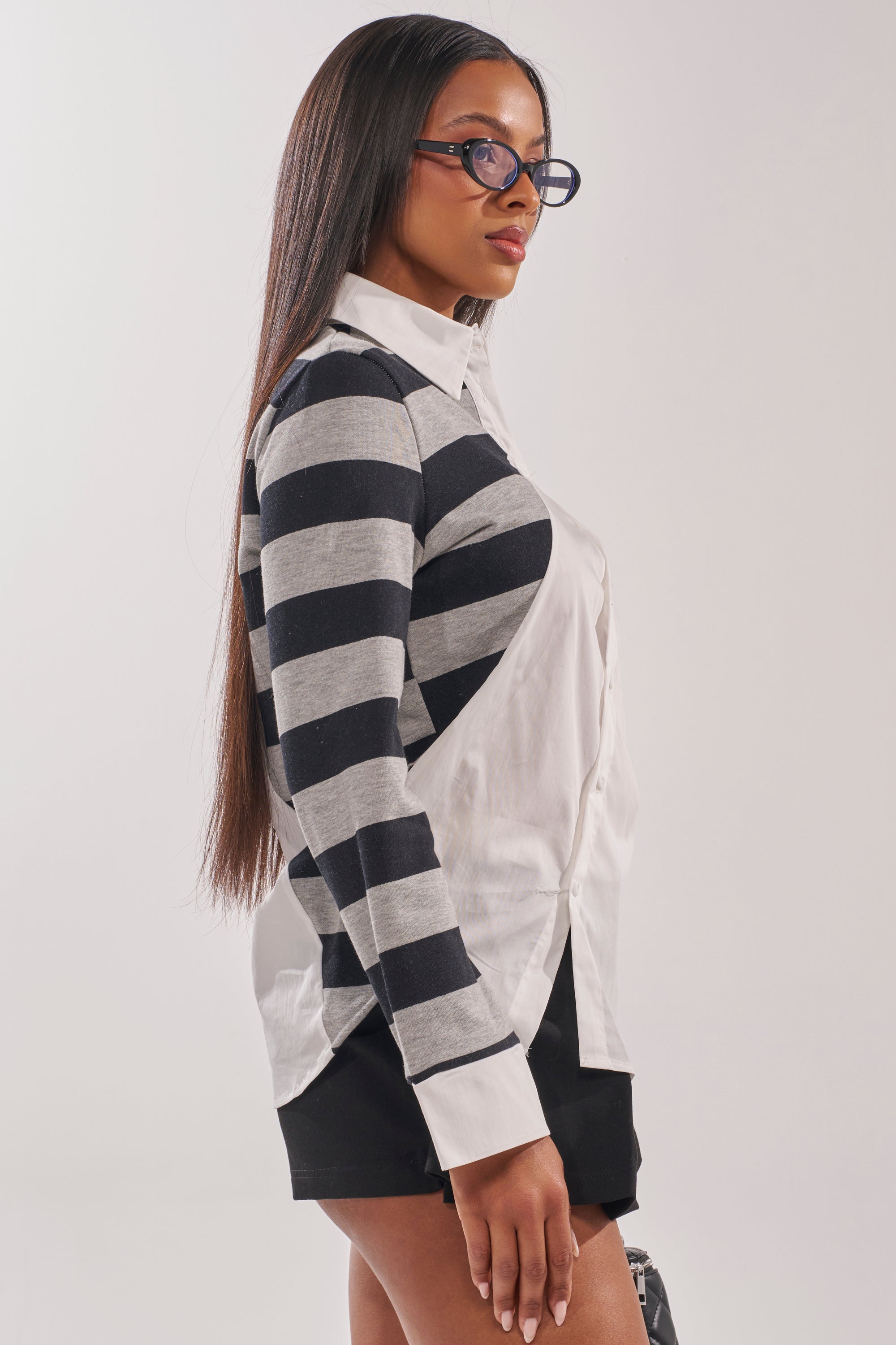 A woman with long straight hair, glasses, and black shorts stands in profile against a plain light background, wearing the MIXED UP STRIPED POLO BLOUSE in black and gray stripes over a crisp white shirt. 3