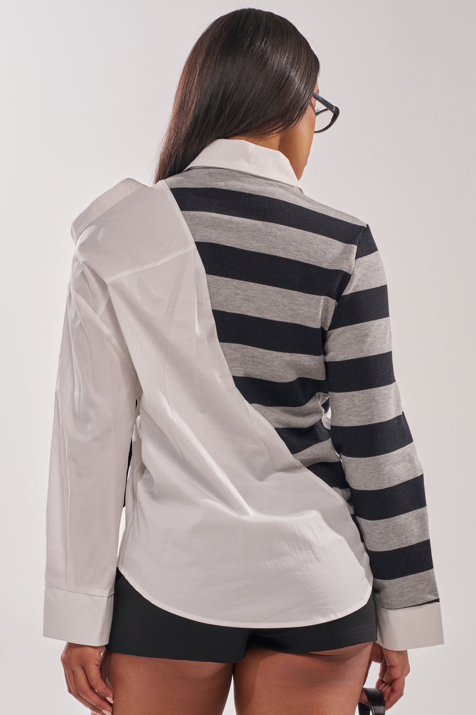 A person with long, straight hair faces away from the camera wearing the MIXED UP STRIPED POLO BLOUSE—half solid white, half black and gray stripes—paired with black shorts. 4