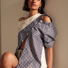 A woman with short dark hair sits on a bench, wearing the MIXED UP TSHIRT BLOUSE over a white top and shorts. She looks slightly upward with a thoughtful expression, softly lit.
