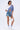 A woman poses in the AKIRA Label MIXED UP TSHIRT BLOUSE—an asymmetrical top with white and blue stripes—styled with denim drawstring shorts, white socks with blue stripes, white heeled boots, and a chic white handbag.