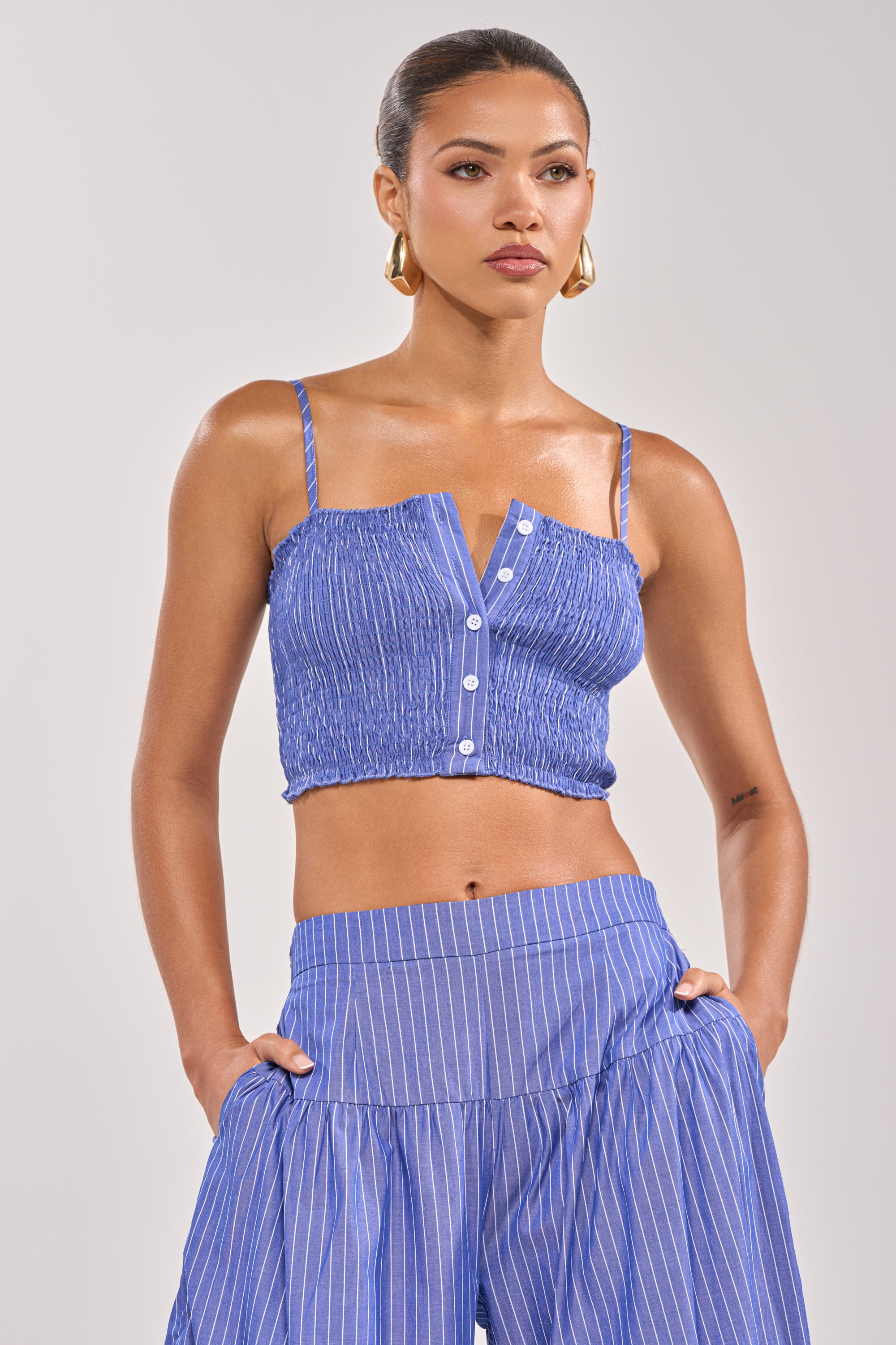 A woman wears the BUSINESS ONLY BRALETTE, a blue pinstriped cotton tank crop top with thin straps and front buttons, paired with matching high-waisted, wide-leg pants, gold hoop earrings, and her hair tied back.