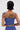 A person with short, dark, wet-look hair is shown from the back wearing the BUSINESS ONLY BRALETTE in blue cotton. Several tattoos decorate their arms and back, including a detailed portrait on the upper back.