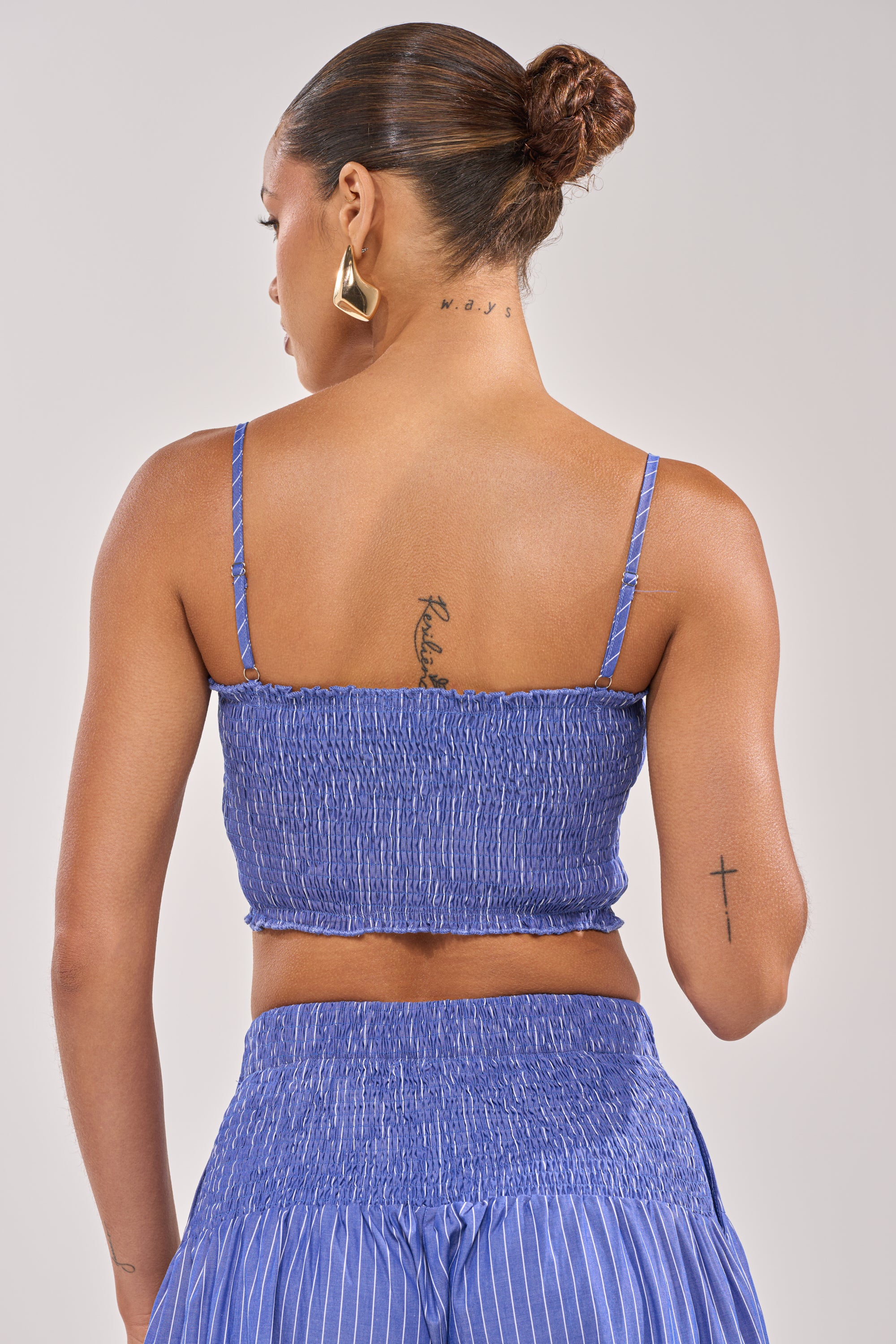 A woman with her hair in a bun and visible neck, upper back, and right arm tattoos faces away from the camera wearing gold geometric earrings and the BUSINESS ONLY BRALETTE paired with a matching blue skirt.