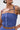 Someone wearing the BUSINESS ONLY BRALETTE, a blue striped cotton crop top with thin straps and buttons. They have short hair, large white earrings, and tattoos visible on their shoulder and torso.