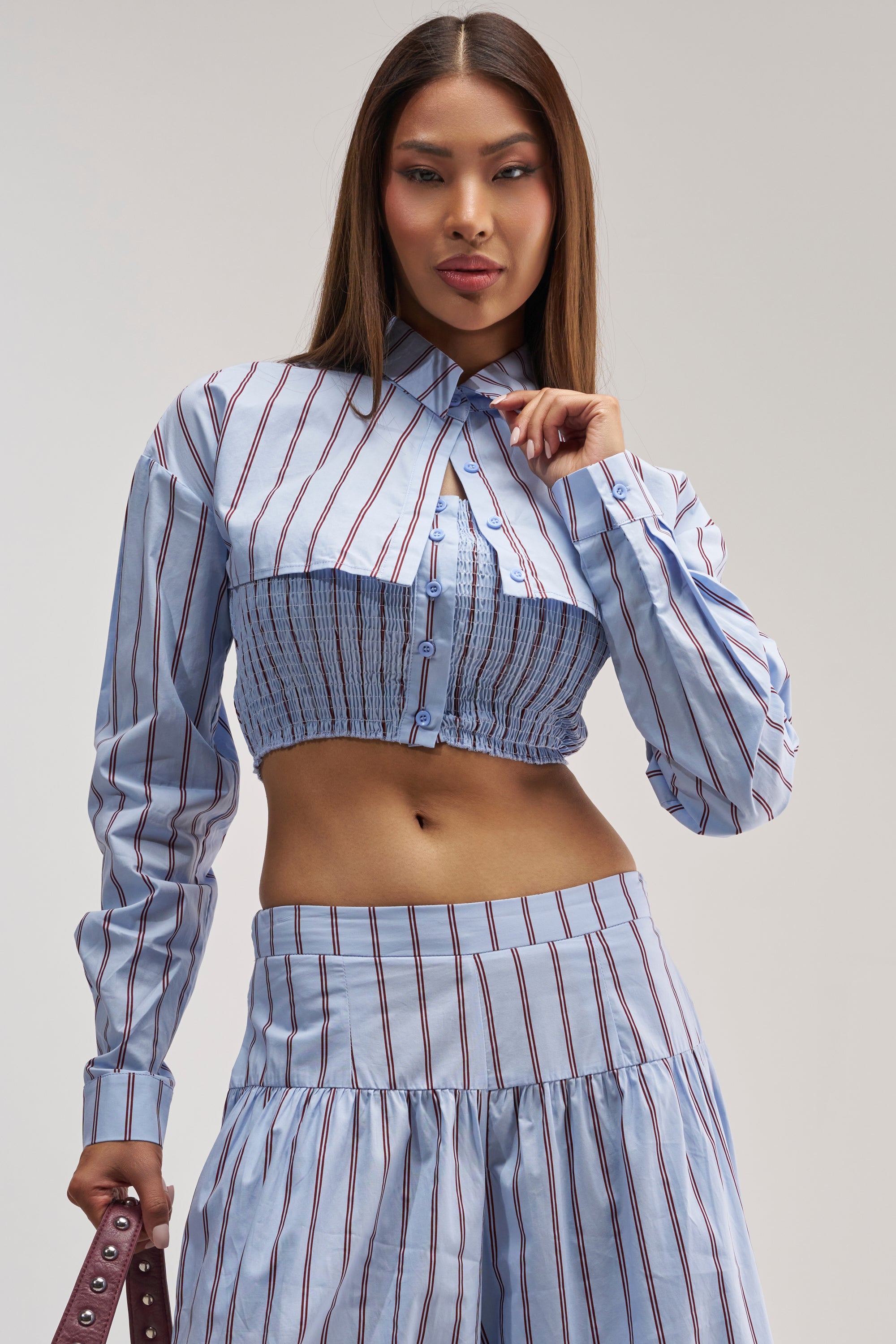 A woman wears the BUSINESS ONLY BUTTON DOWN IN LIGHT BLUE—a light blue pinstripe, long-sleeve cropped button down—paired with a matching high-waisted skirt and poses confidently against a plain background, making it the perfect chic summer date night top.