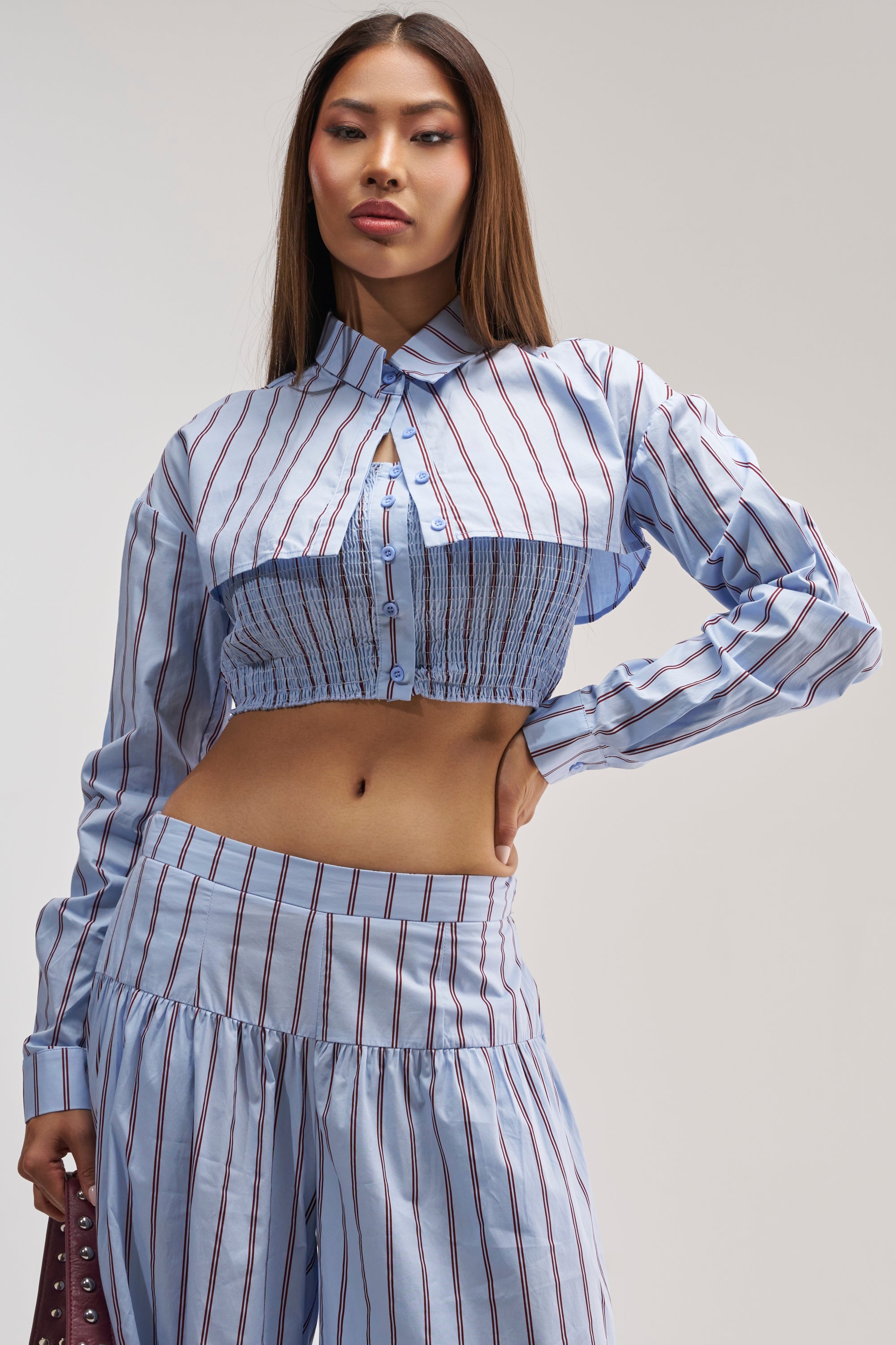 A woman wearing the BUSINESS ONLY BUTTON DOWN IN LIGHT BLUE—a light blue pinstripe blouse with long sleeves, ruching, and cropped button-down styling—paired with matching high-waisted pants, stands against a light background for an effortlessly chic summer date night look.