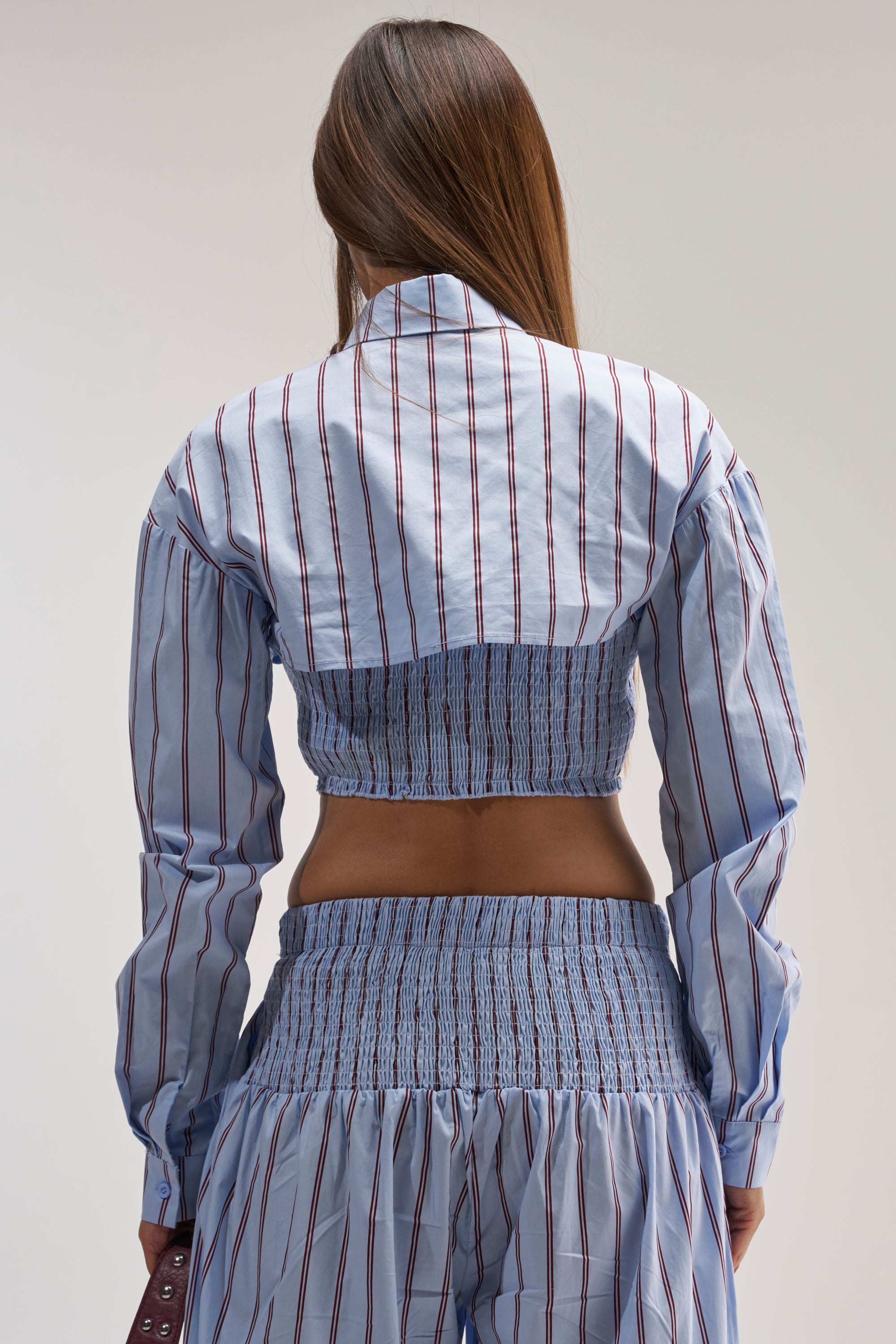 A woman with long brown hair is seen from the back against a plain background, wearing the BUSINESS ONLY BUTTON DOWN IN LIGHT BLUE styled as a smocked crop top with matching high-waisted pants.