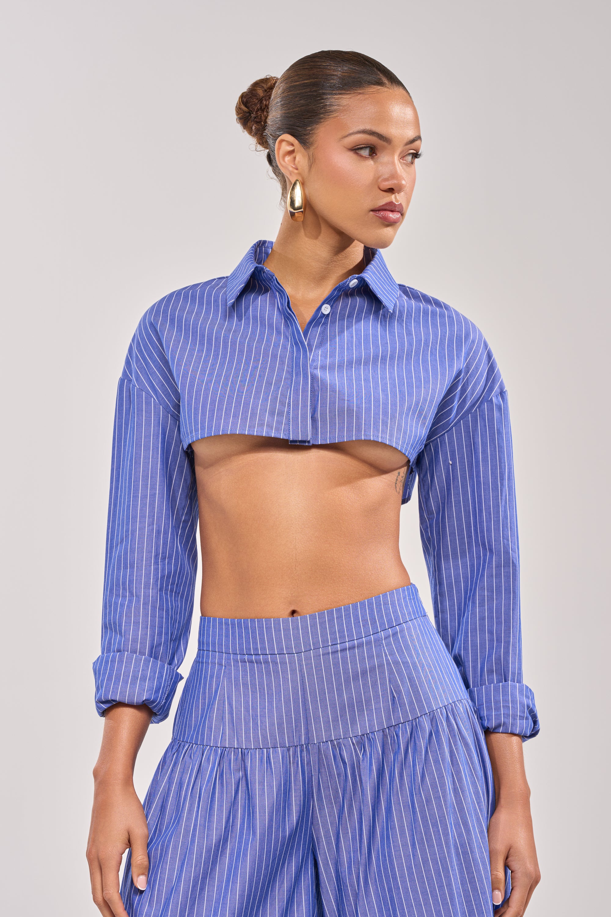 A woman wears the BUSINESS ONLY BUTTON DOWN, a blue-striped, cropped shirt with high-waisted pants. Her hair is in a bun and she has gold hoop earrings, standing against a plain light background and gazing to the side.