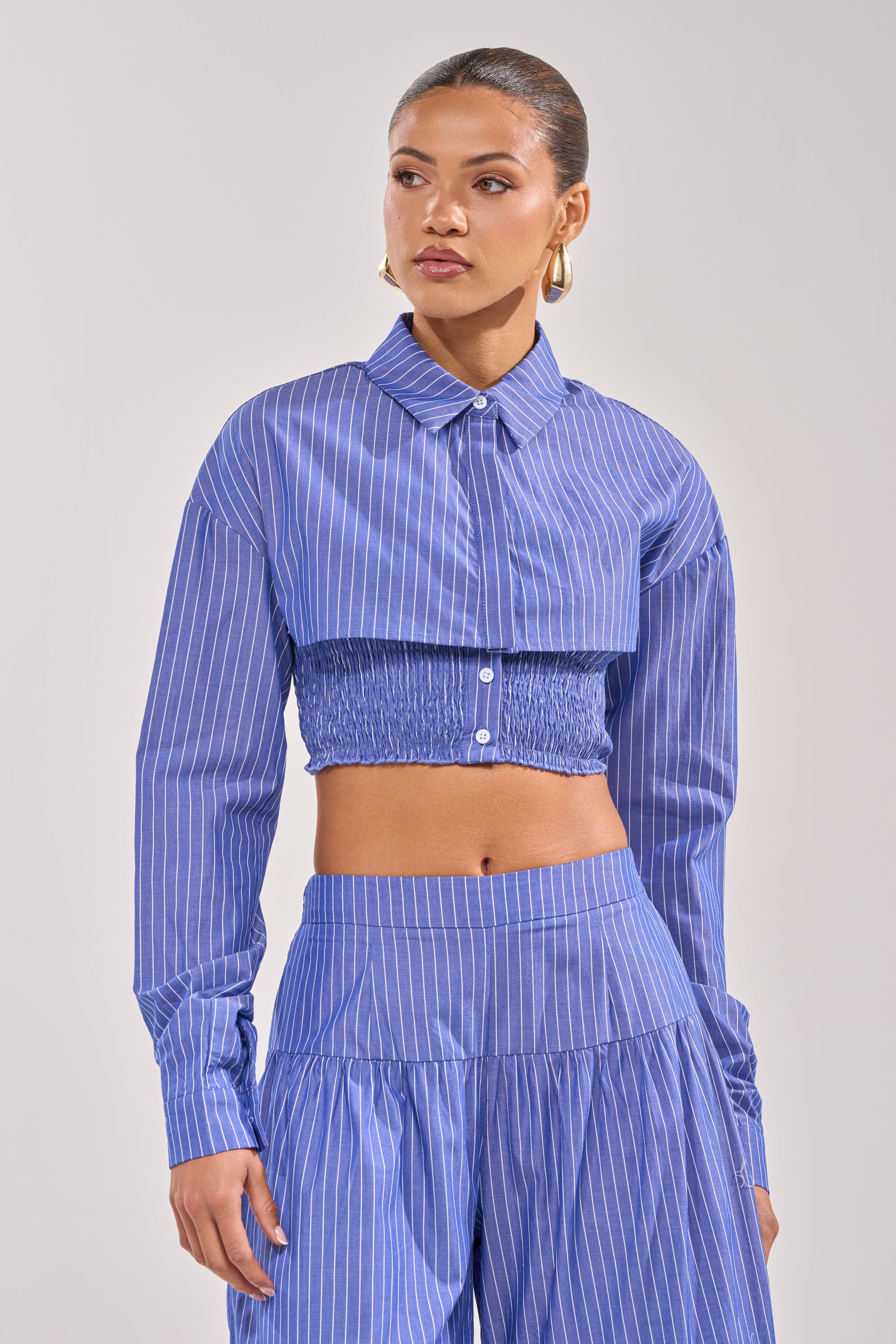A woman wears the BUSINESS ONLY BUTTON DOWN—a blue-striped, cropped silhouette—paired with matching high-waisted, wide-leg pants, standing against a plain background and gazing slightly to the side.