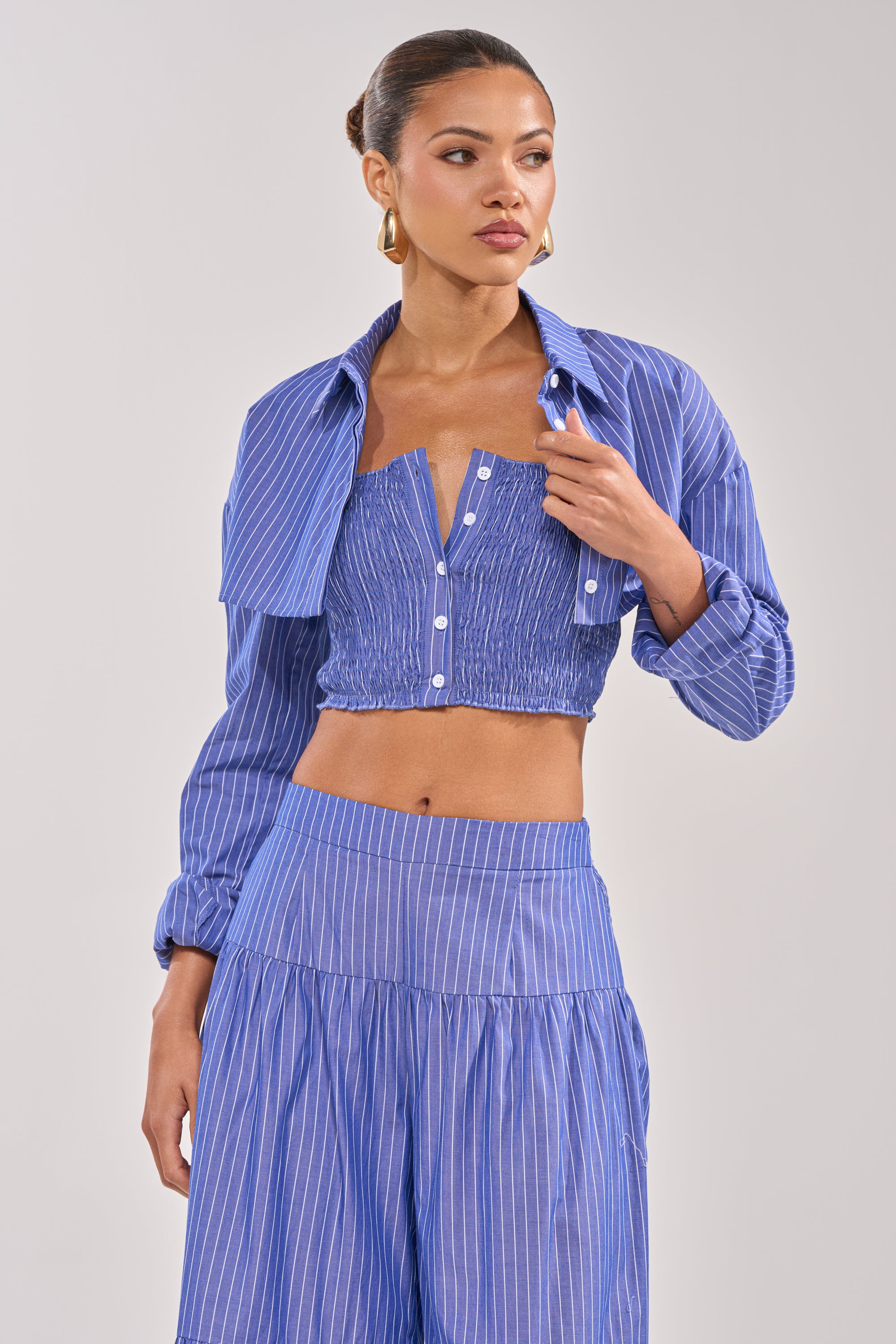 A woman styles the BUSINESS ONLY BUTTON DOWN set—blue-striped, cropped shirt and high-waisted flowy pants—with her hair up and gold hoop earrings, glancing slightly to the side.