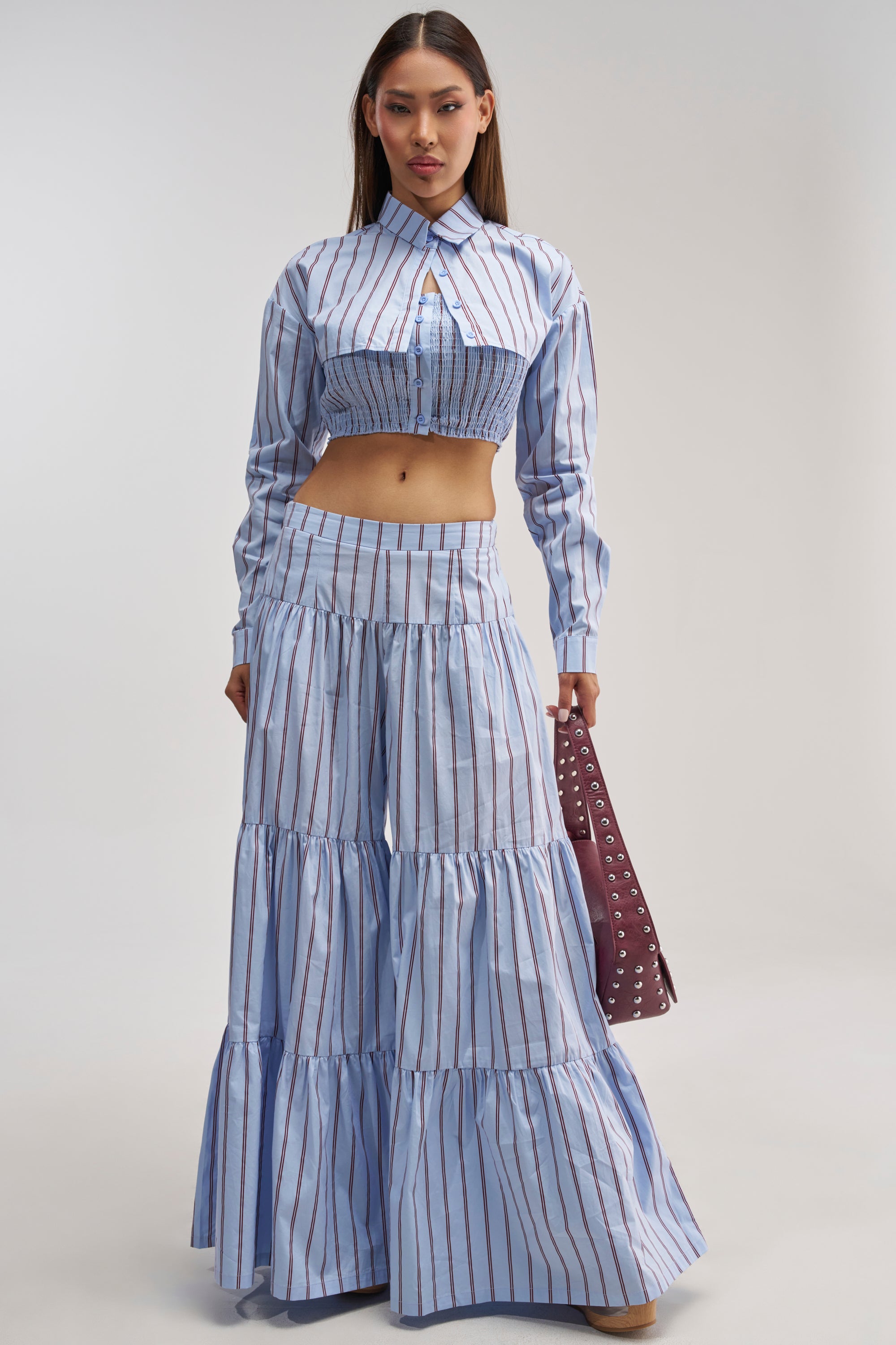 A woman stands against a plain background wearing the Business Only Stripe Palazzo Pant in Light Blue—a high-rise, wide-leg pant with tiered ruffle legs—paired with a matching blue and white striped crop top and holding a purple bag.
