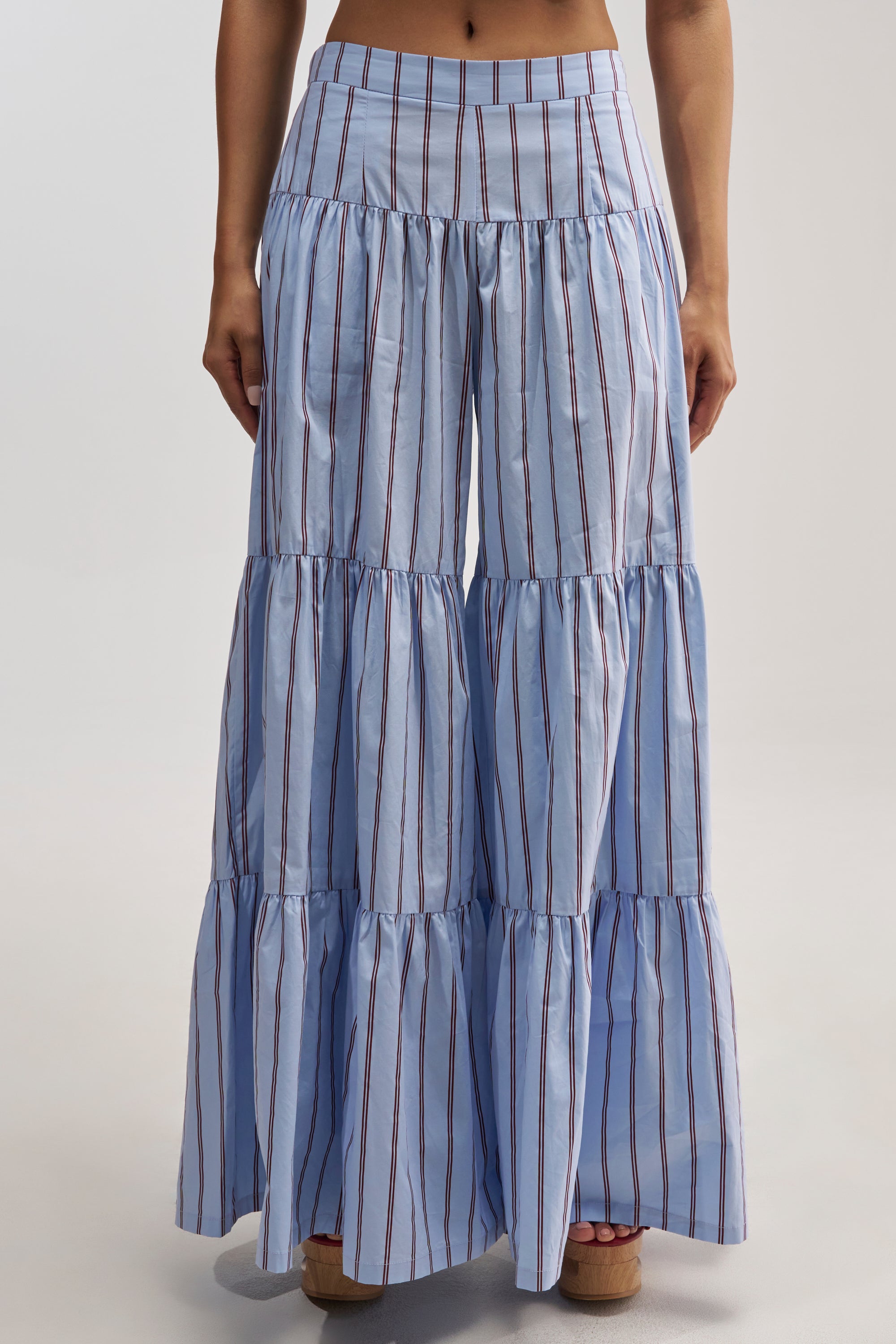 A person wearing the BUSINESS ONLY STRIPE PALAZZO PANT IN LIGHT BLUE, which features high-rise wide-leg styling, light blue fabric with vertical red and white stripes, and tiered ruffle legs, stands against a plain background.
