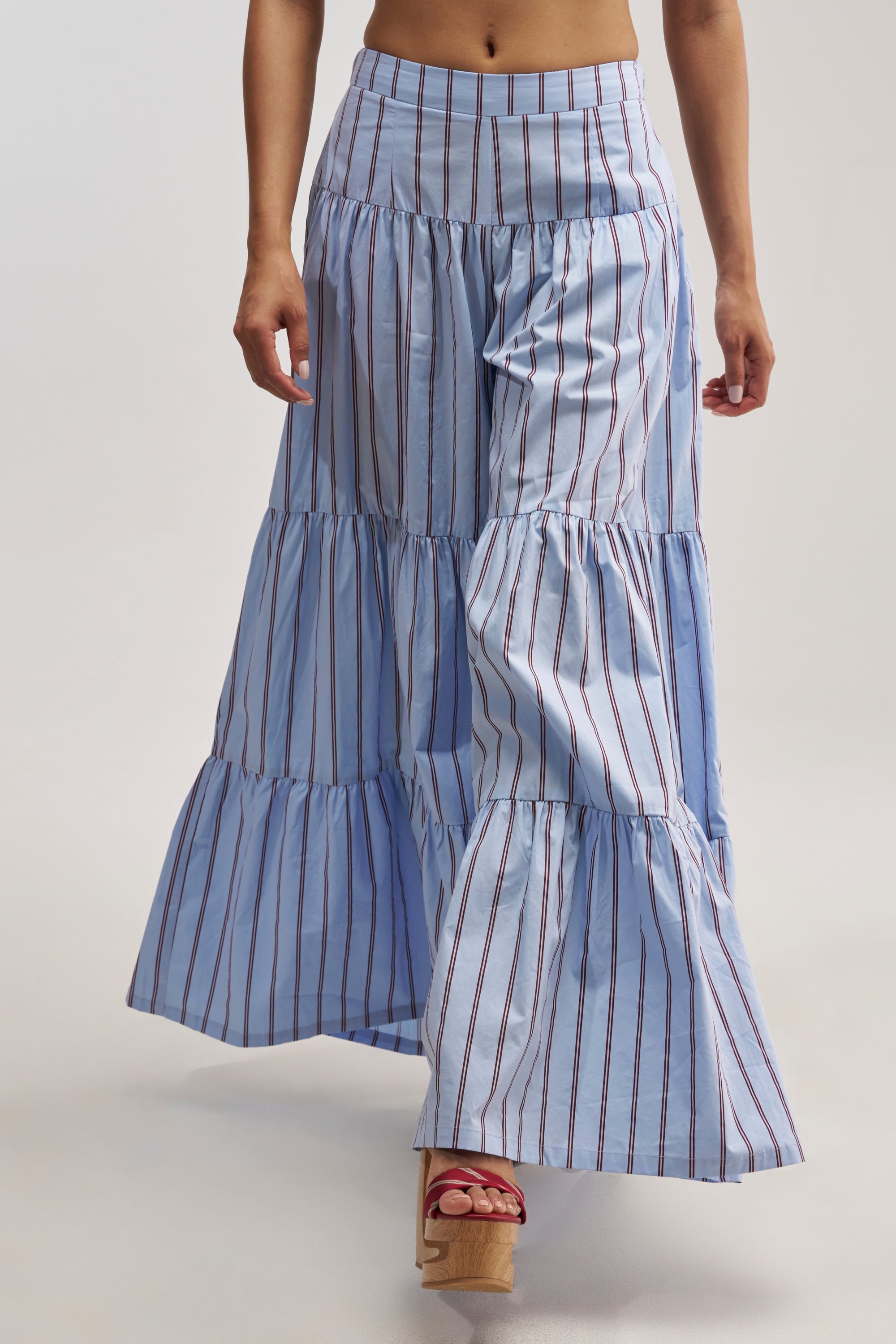 A person wearing the BUSINESS ONLY STRIPE PALAZZO PANT IN LIGHT BLUE, featuring long, tiered, blue fabric with vertical red stripes reminiscent of ruffle pants, paired with red wedge sandals. Only the lower half of their body is visible against a plain background.
