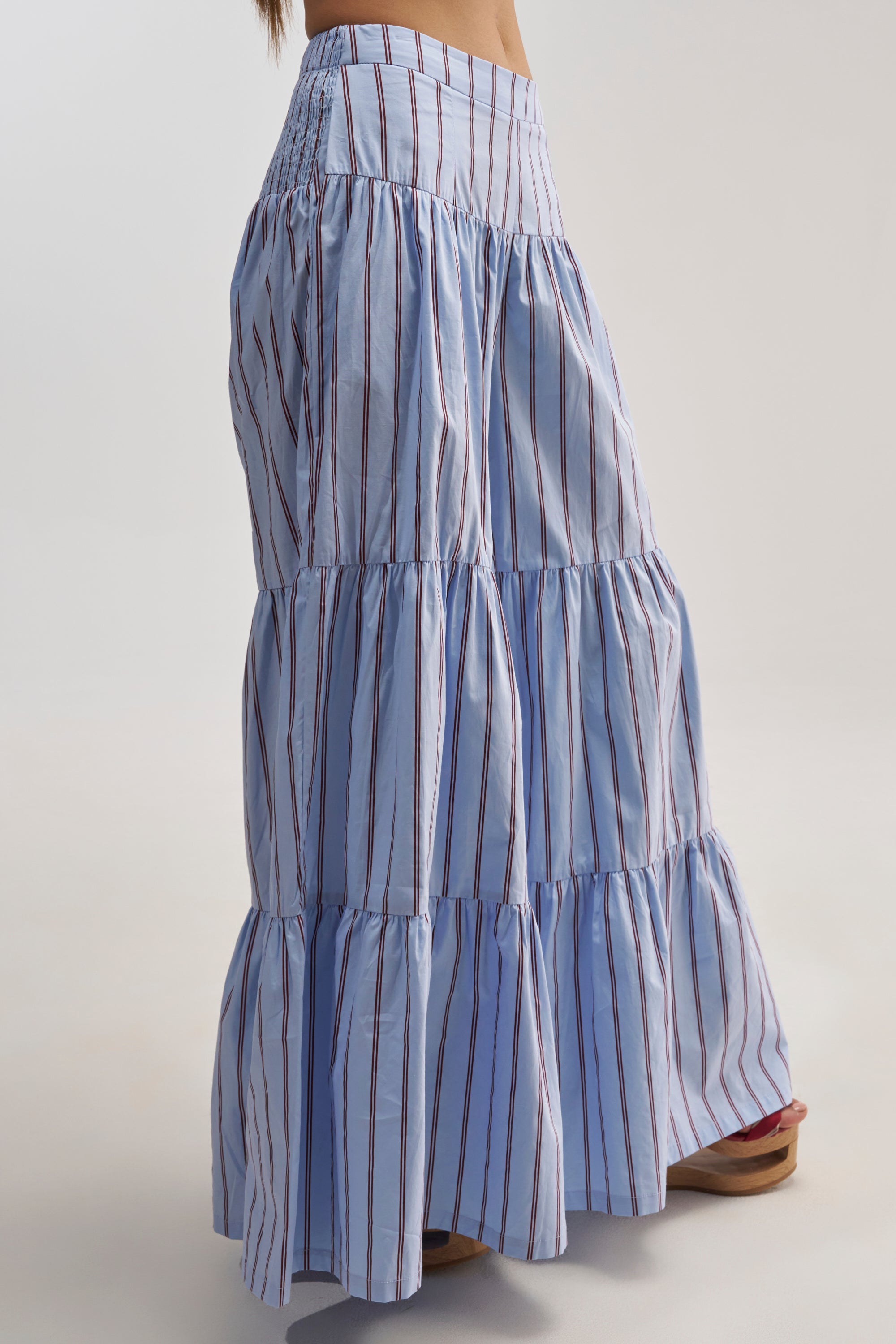 A person wearing the BUSINESS ONLY STRIPE PALAZZO PANT IN LIGHT BLUE, featuring long, tiered styling with vertical red and white pinstripes, paired with brown sandals, stands against a plain background.