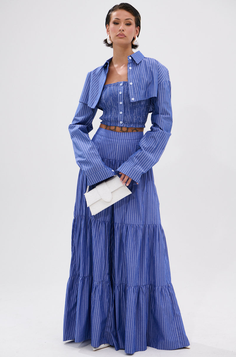 A woman stands against a plain white background wearing the BUSINESS ONLY STRIPE PALAZZO PANT with blue and white stripes, paired with a matching crop top, cropped jacket, and holding a small white clutch.