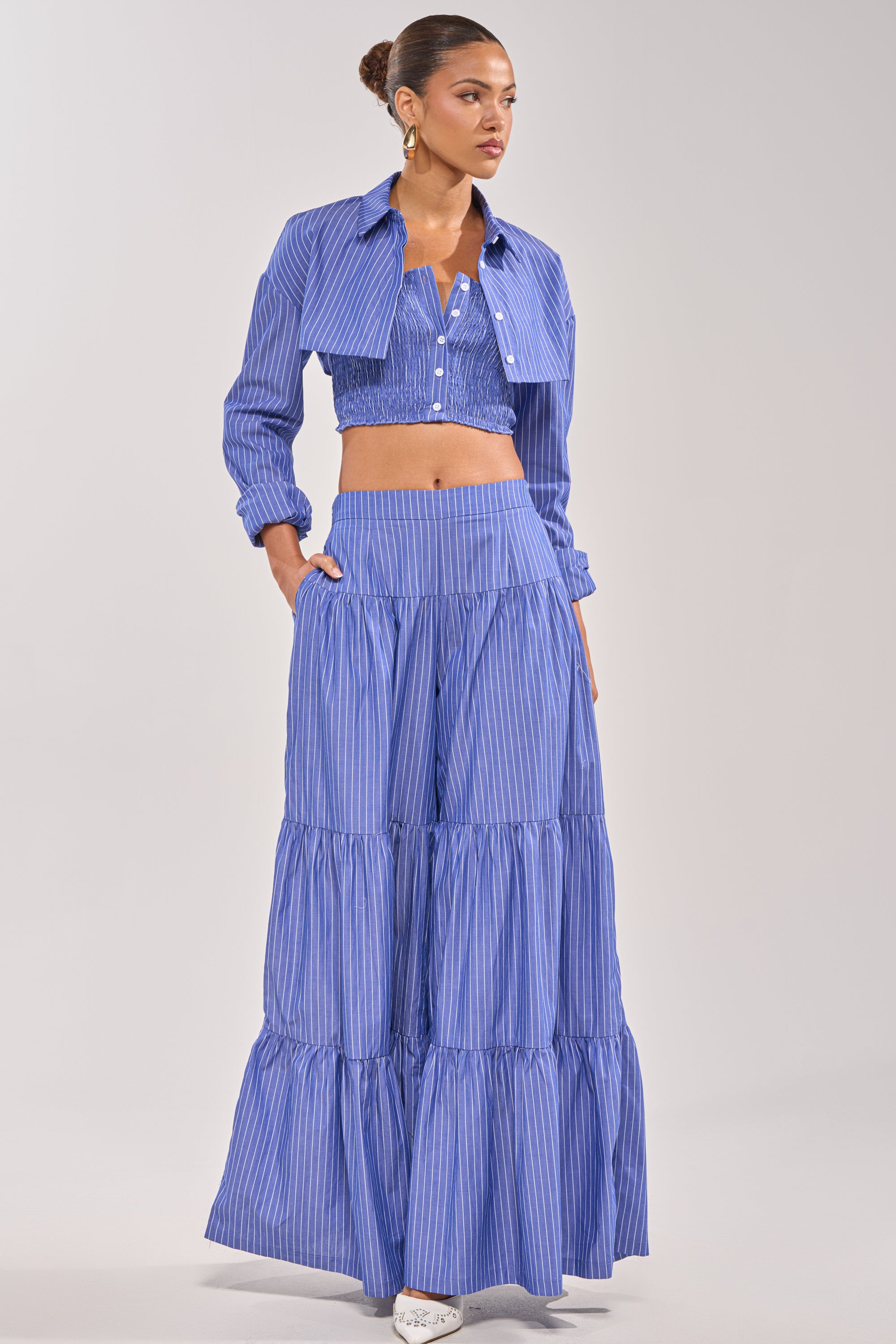 A woman wears the BUSINESS ONLY STRIPE PALAZZO PANT, a blue-and-white pinstriped style featuring a high waist and wide legs—an effortless summer look. She stands confidently with her hands in the pockets.
