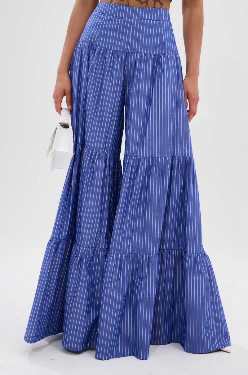 A person wears the BUSINESS ONLY STRIPE PALAZZO PANT—a high-rise, wide-leg blue pant with white stripes—styled with a brown top and a small white handbag.
