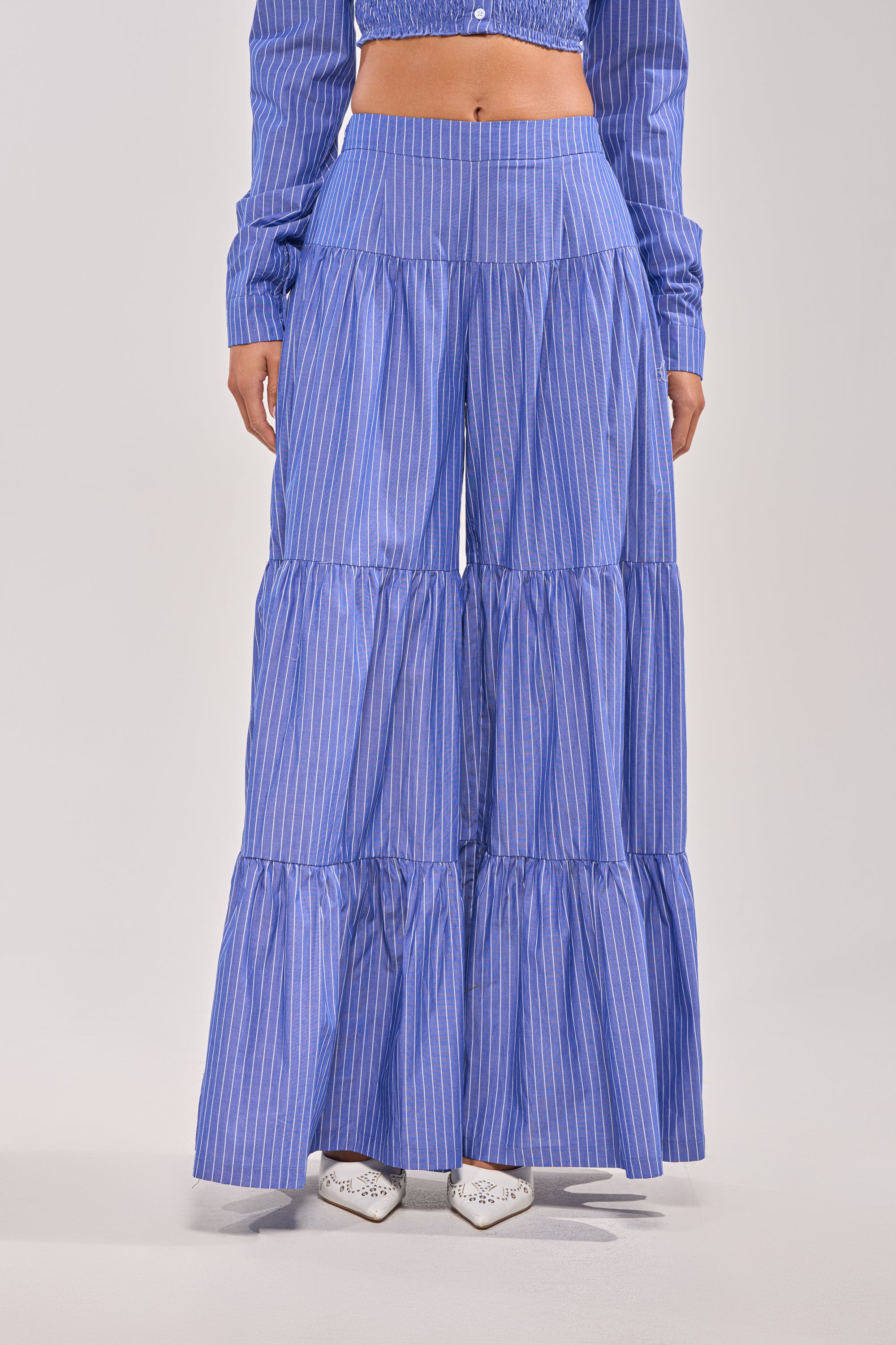 Wearing the BUSINESS ONLY STRIPE PALAZZO PANT, a high-rise, wide-leg blue striped design with three ruffled tiers, paired with white pointed shoes and a matching long-sleeved top for a statement look.