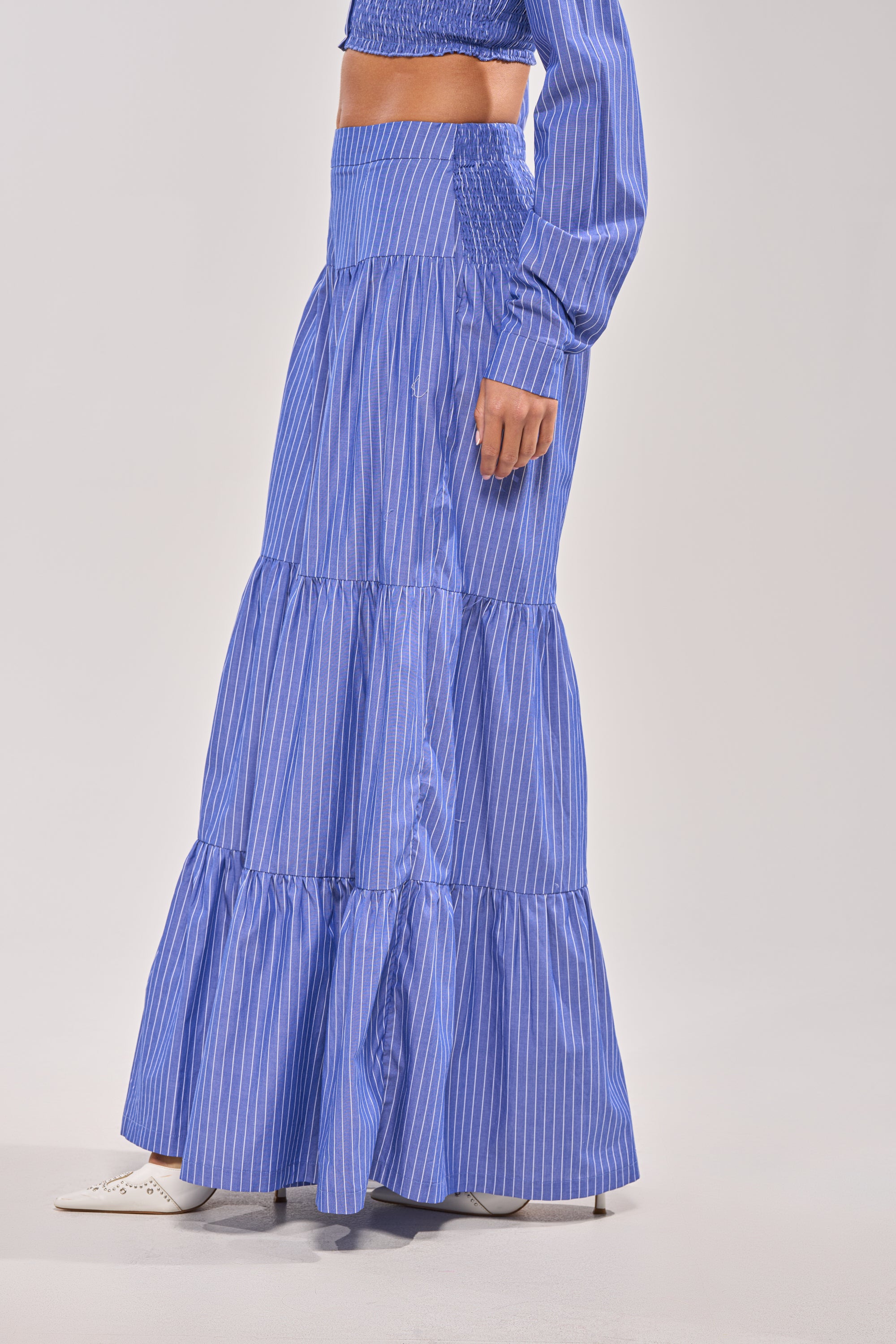 A person stands against a plain background wearing the BUSINESS ONLY STRIPE PALAZZO PANT in blue and white vertical stripes, paired with a matching long-sleeve crop top and white shoes.