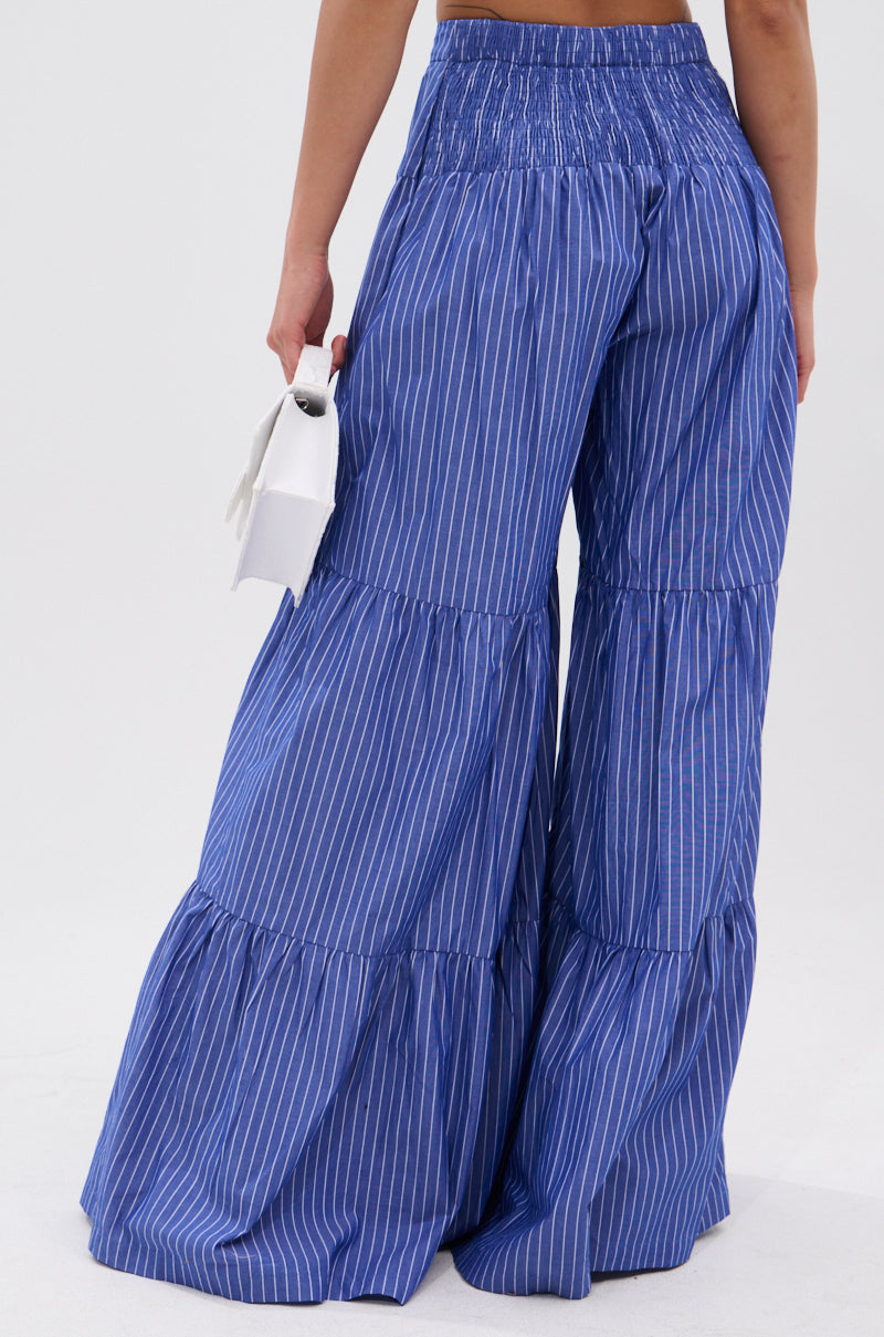 A person stands facing away, wearing the BUSINESS ONLY STRIPE PALAZZO PANT featuring a high-rise fit and wide-leg silhouette with blue and white vertical stripes and tiered ruffles, holding a small white handbag against a light background.