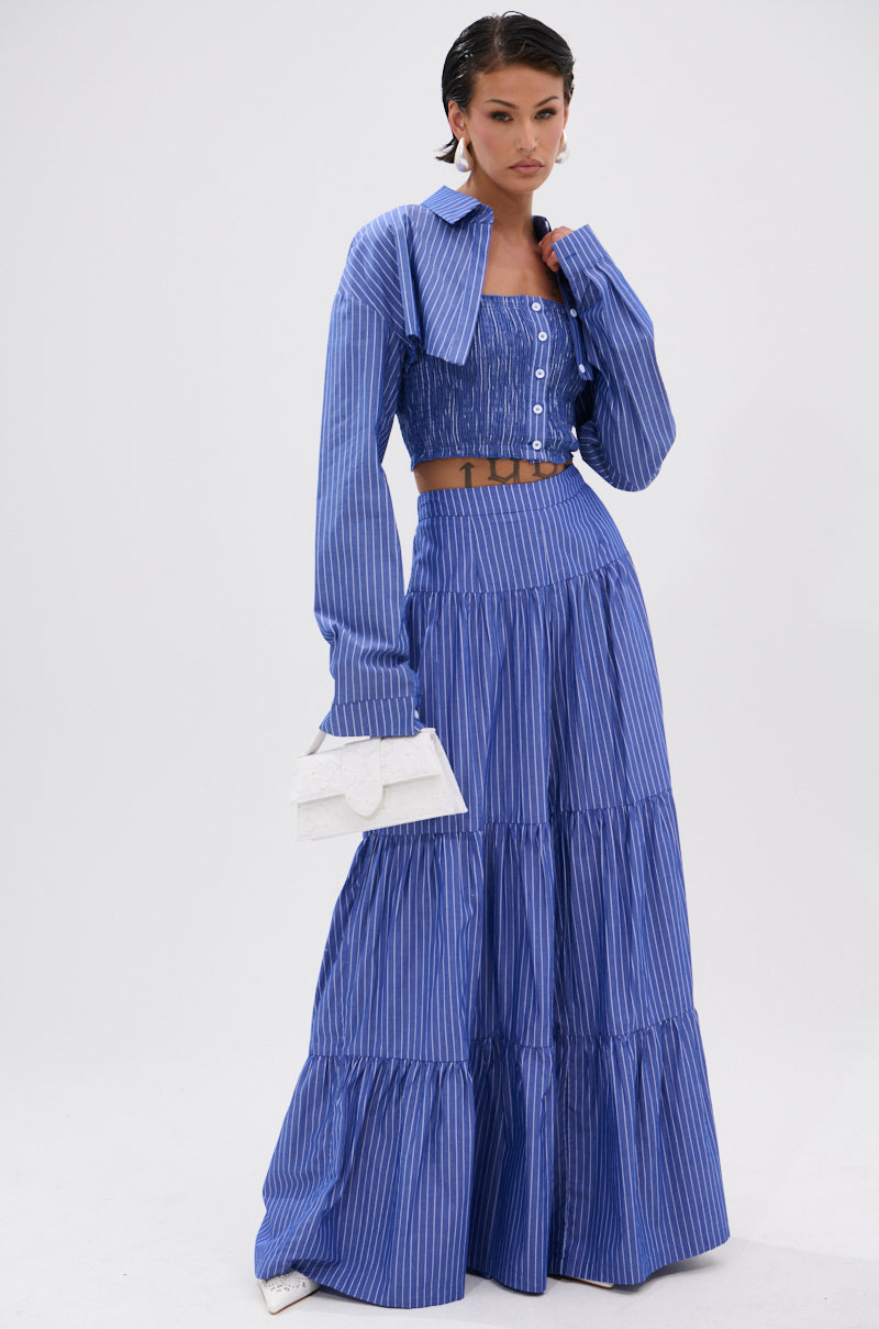 A woman stands against a plain white background wearing the BUSINESS ONLY STRIPE PALAZZO PANT—a blue and white striped, high-rise, wide-leg pant—paired with a matching cropped jacket, smocked button-up top, and holding a white clutch.
