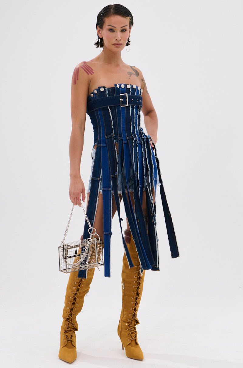 A woman in the GOT A LOT GOING ON DENIM CORSET dress, tan lace-up knee-high boots, and a clear box-shaped handbag stands against a white background, showcasing upcycled fashion with slicked-back hair and arm tattoos. 0