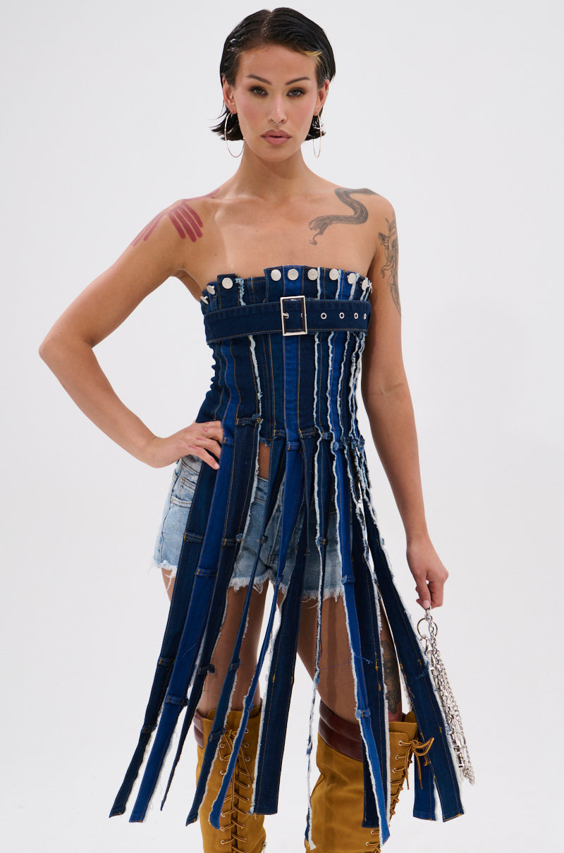 A person confidently stands against a plain white background wearing the GOT A LOT GOING ON DENIM CORSET, paired with distressed denim shorts, brown knee-high boots, large hoop earrings, and holding a chain handbag. 1