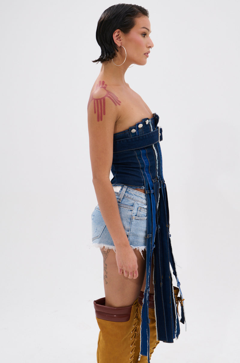 A woman with wet-look, short dark hair wears large hoop earrings, tall brown lace-up boots, light denim shorts, and the GOT A LOT GOING ON DENIM CORSET dress with frayed edges. She has a pink painted shoulder and stands in profile. 3