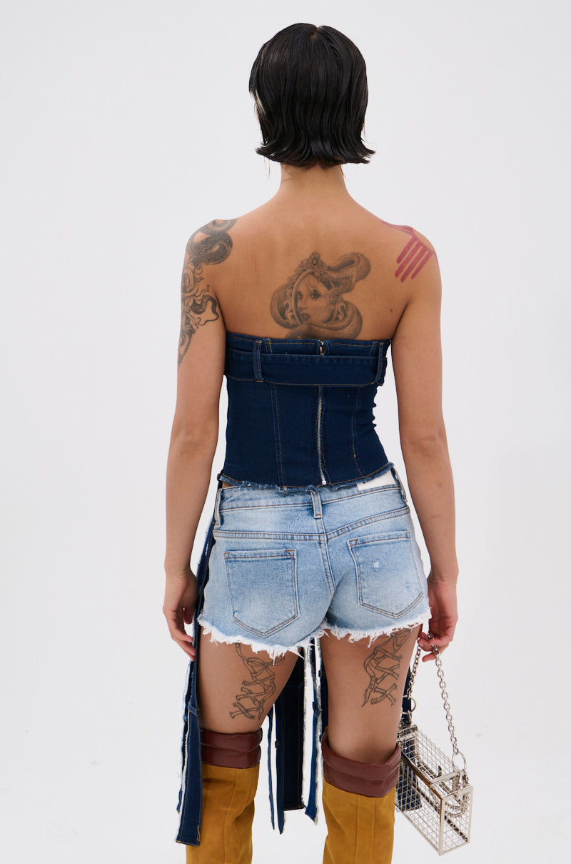 A person with short dark hair and tattoos models the GOT A LOT GOING ON DENIM CORSET, paired with distressed shorts, tan boots, and a small square chain-strap handbag, facing away from the camera. 4
