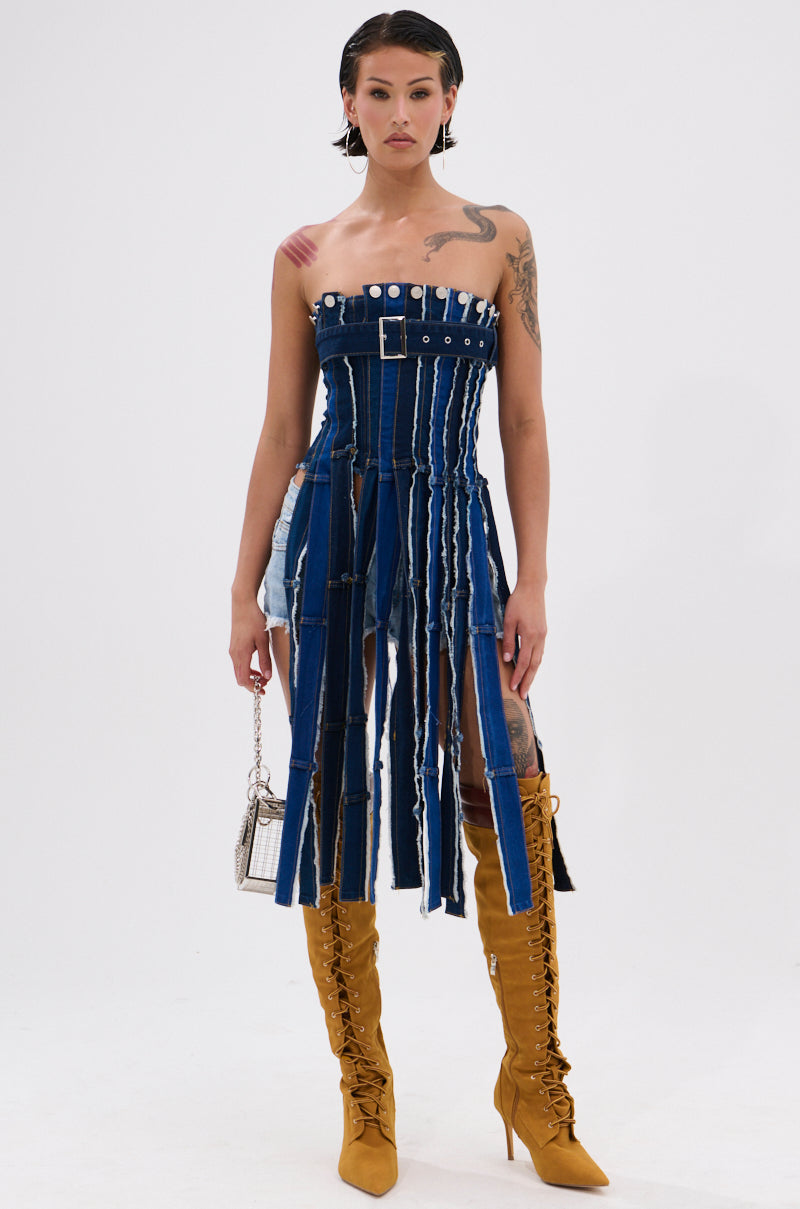 Wearing the GOT A LOT GOING ON DENIM CORSET dress with vertical strips and a belt, a tattooed person pairs it with knee-high mustard yellow lace-up boots and a small silver handbag, standing against a plain white background. 5